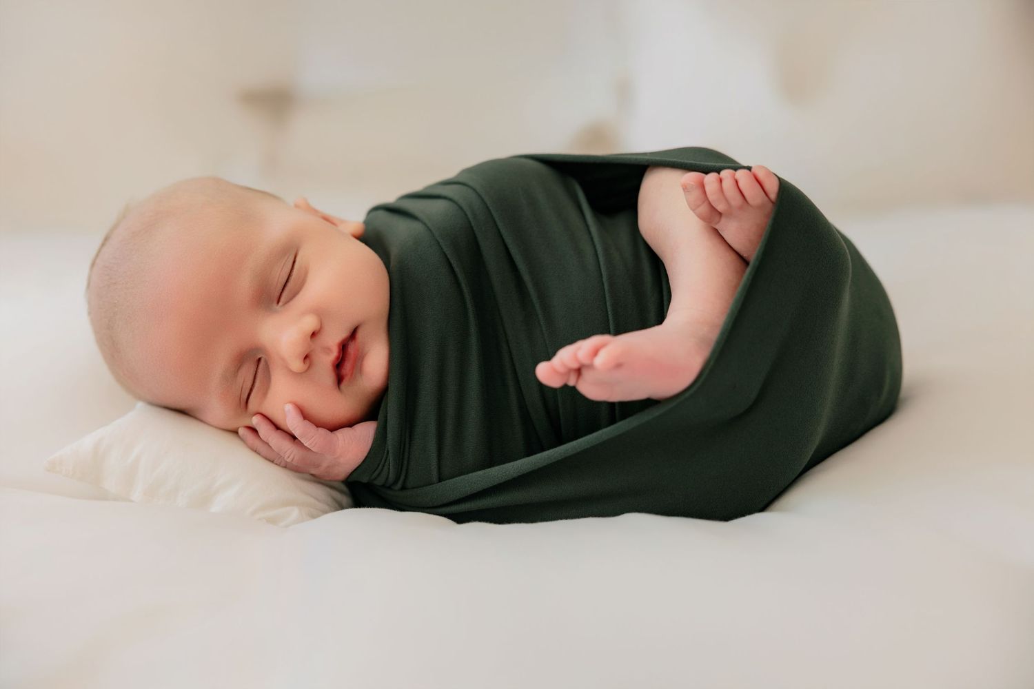 Sleeping newborn baby swaddled in a dark green wrap, resting peacefully on a white surface.