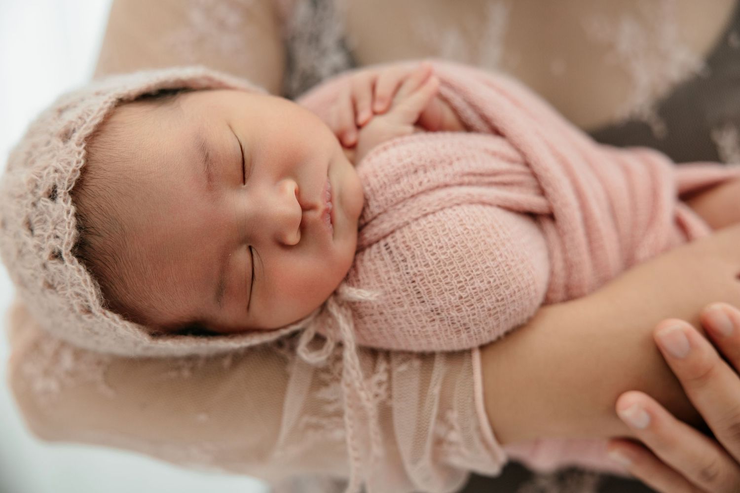 Newborn baby sleeping peacefully wrapped in a soft pink knit blanket and lace bonnet, cradled in loving arms.