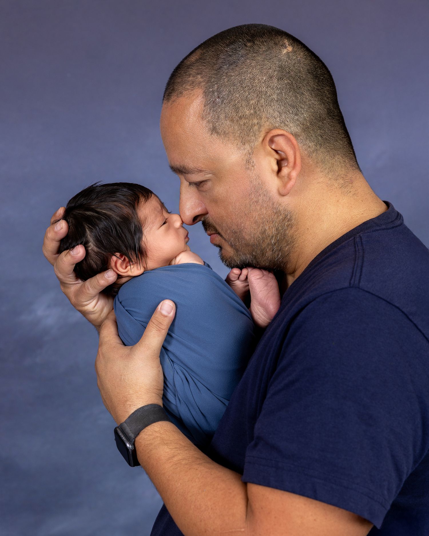 A father tenderly holds his newborn baby close, touching noses in an intimate studio portrait with blue backdrop.