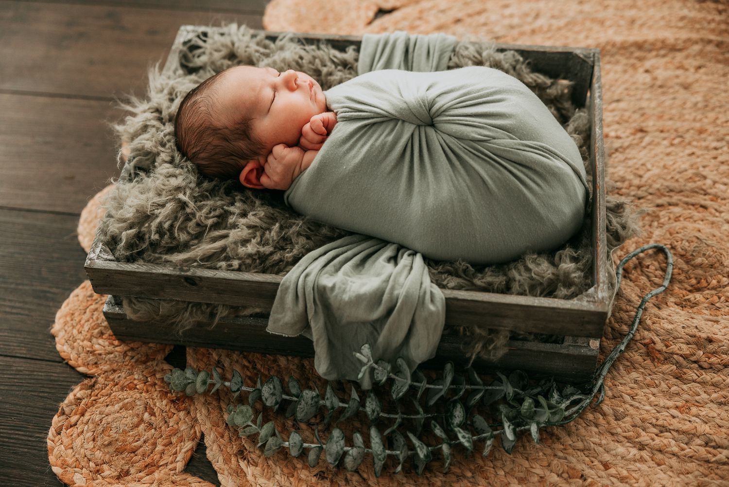 Newborn baby swaddled in sage green wrap sleeping in rustic wooden crate with soft gray fur blanket.