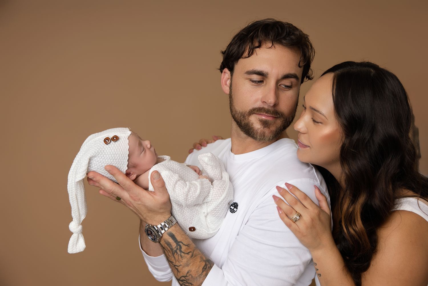 A couple in white shirts tenderly holds their newborn baby wearing a white knit hat against a tan background.
