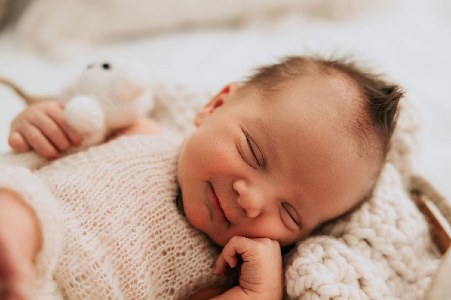 Peaceful smiling newborn baby wrapped in cream knit blanket with tiny teddy bear during newborn photography session.