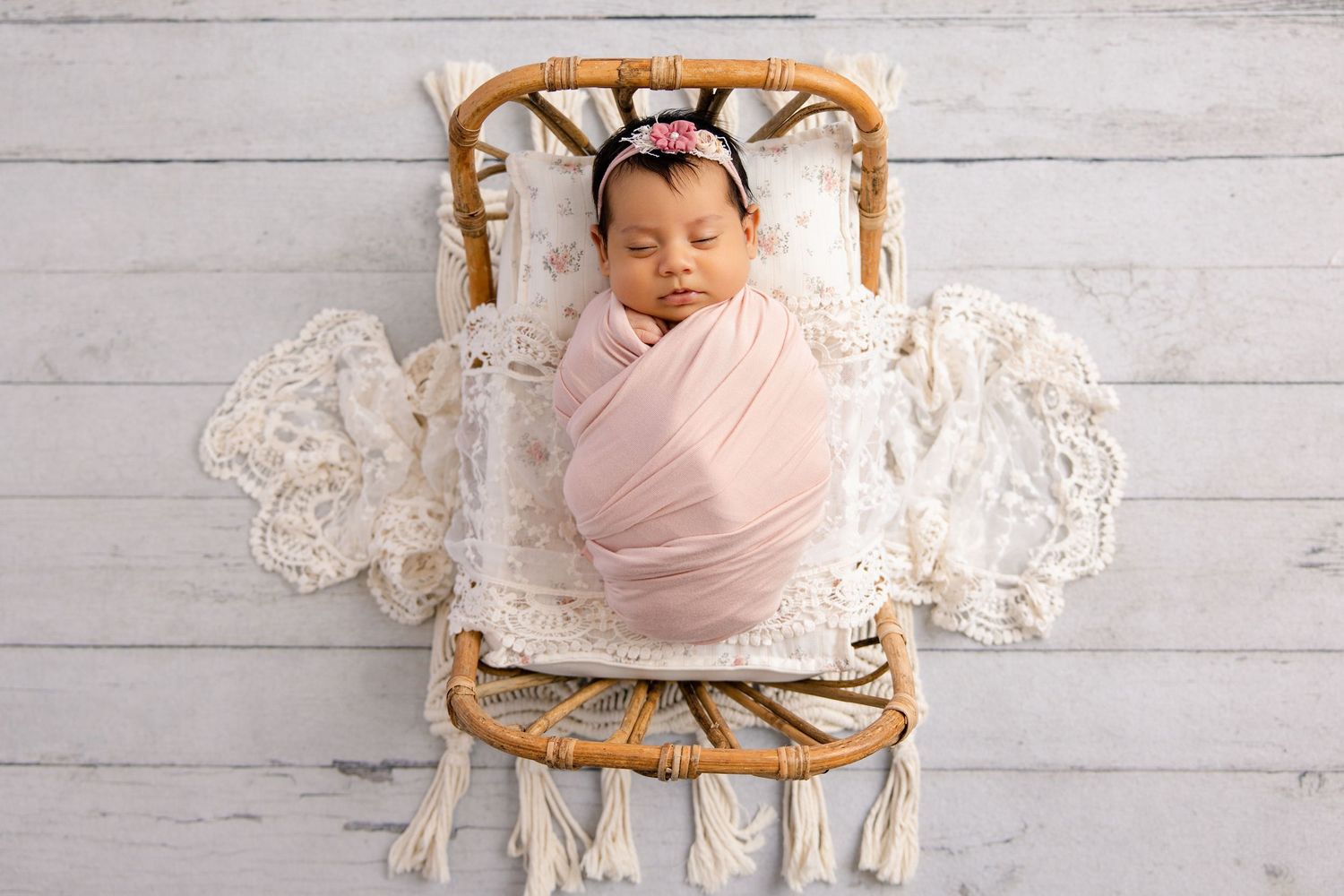 Sleeping newborn baby girl swaddled in pink wrap with floral headband in a rattan bed.