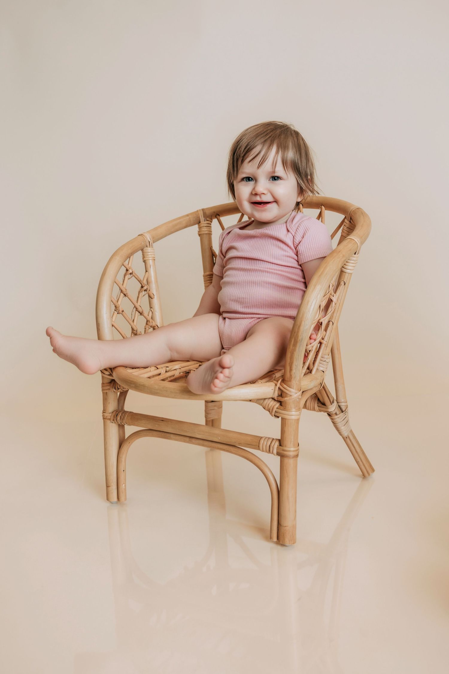 Smiling baby girl in pink bodysuit sitting in a small rattan chair against a beige background.