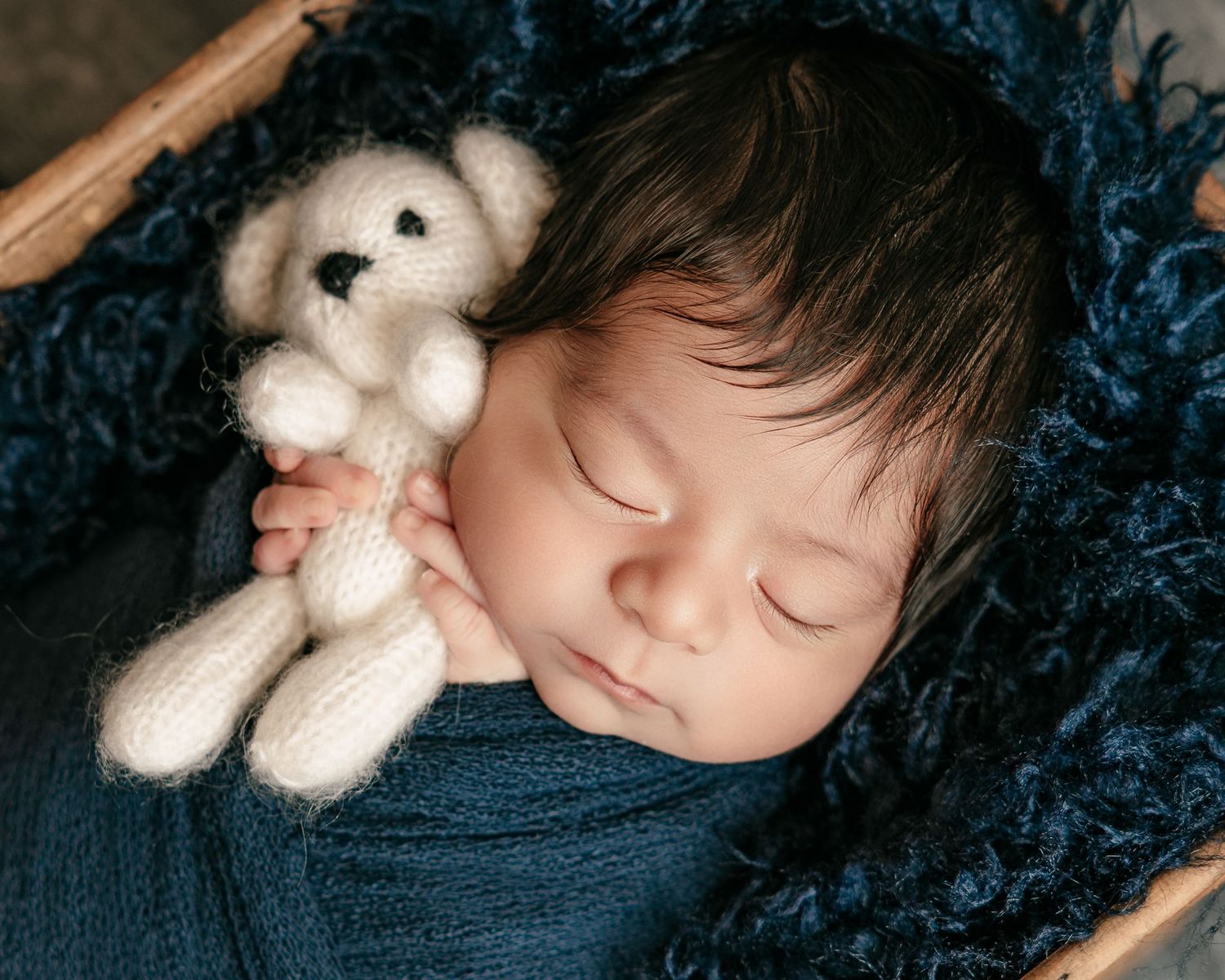 Sleeping newborn baby holding a white knitted teddy bear, wrapped in blue, nestled in a cozy dark blue basket.