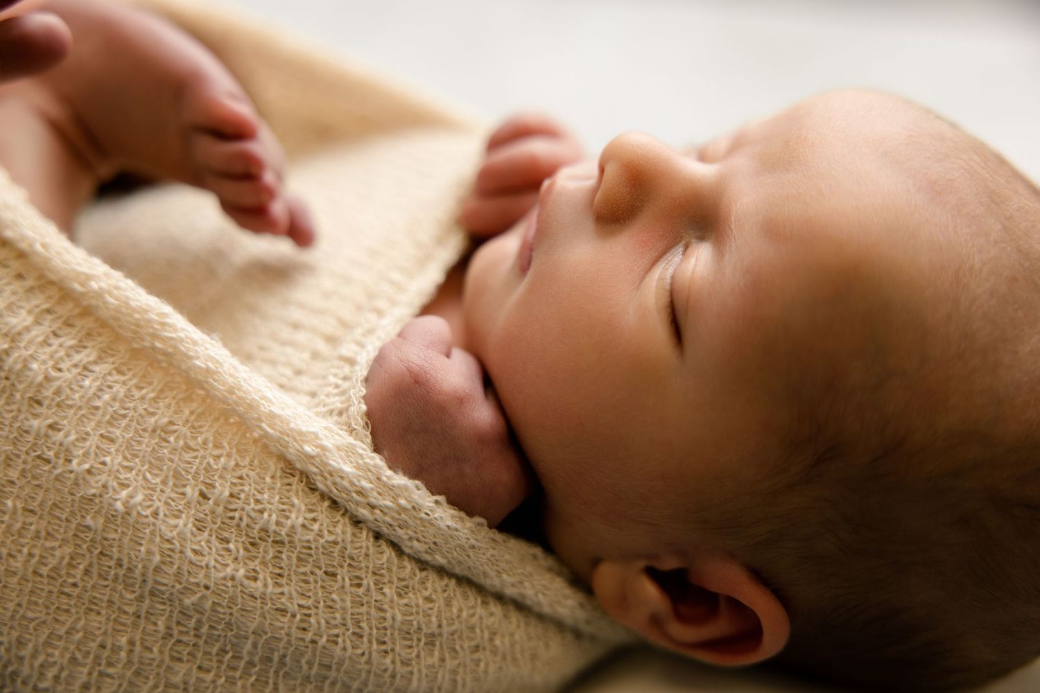 Peaceful newborn baby sleeping soundly, swaddled in a soft cream knit blanket with tiny hands visible.