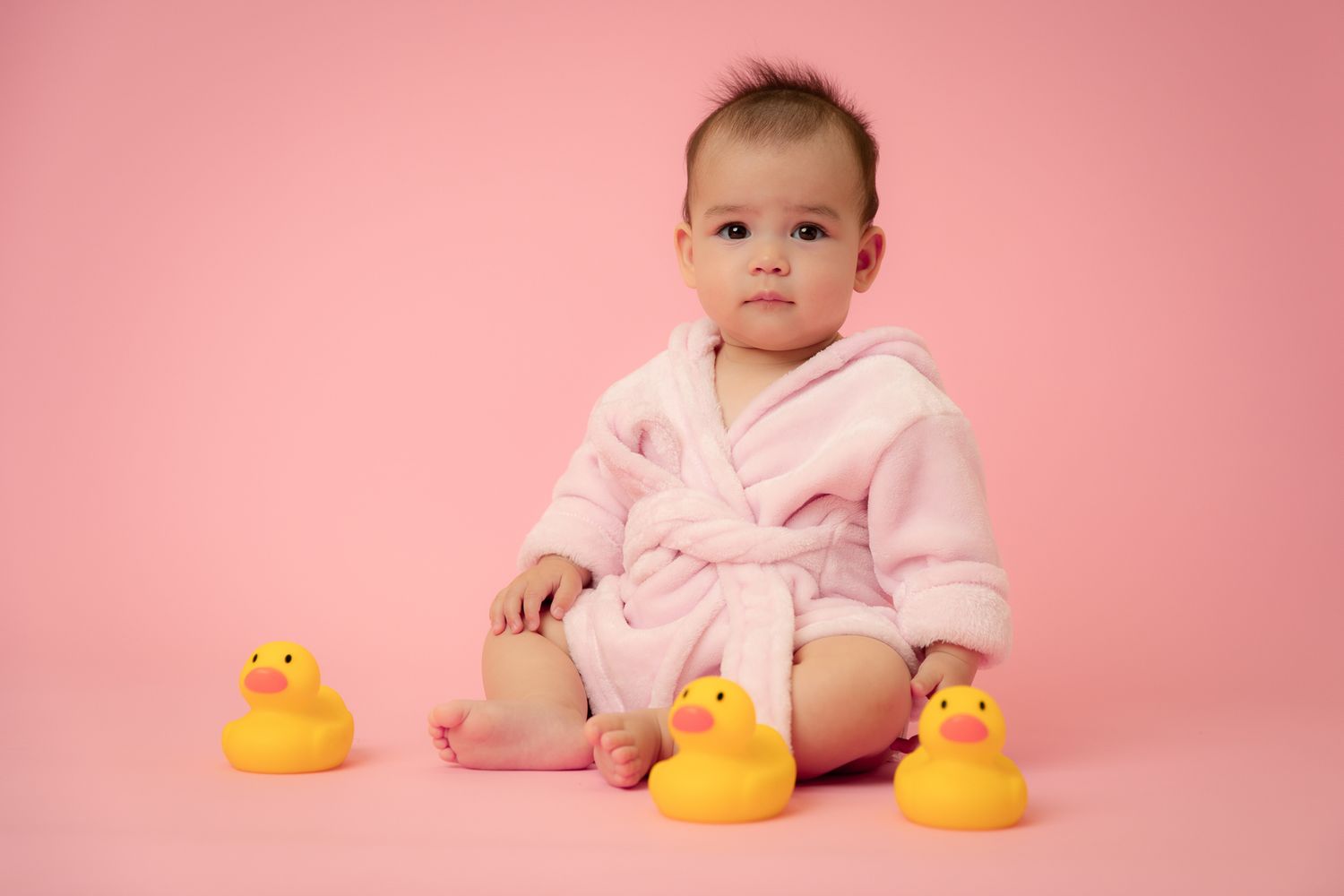 Baby girl in pink robe sitting with rubber ducks on pink background.