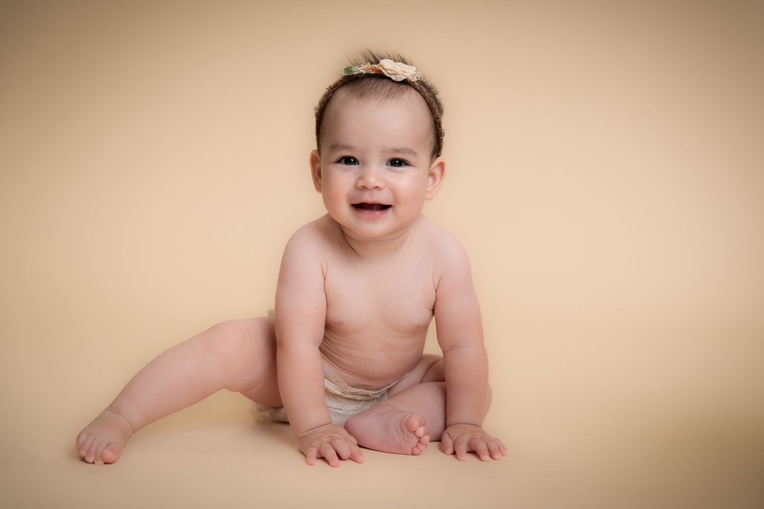 Smiling baby girl in diaper with bow headband sitting on beige backdrop.