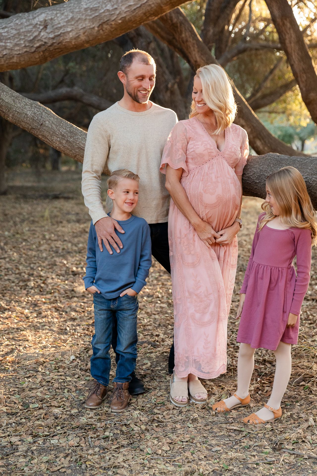 Pregnant woman with family of four poses near leaning trees in warm sunset light.