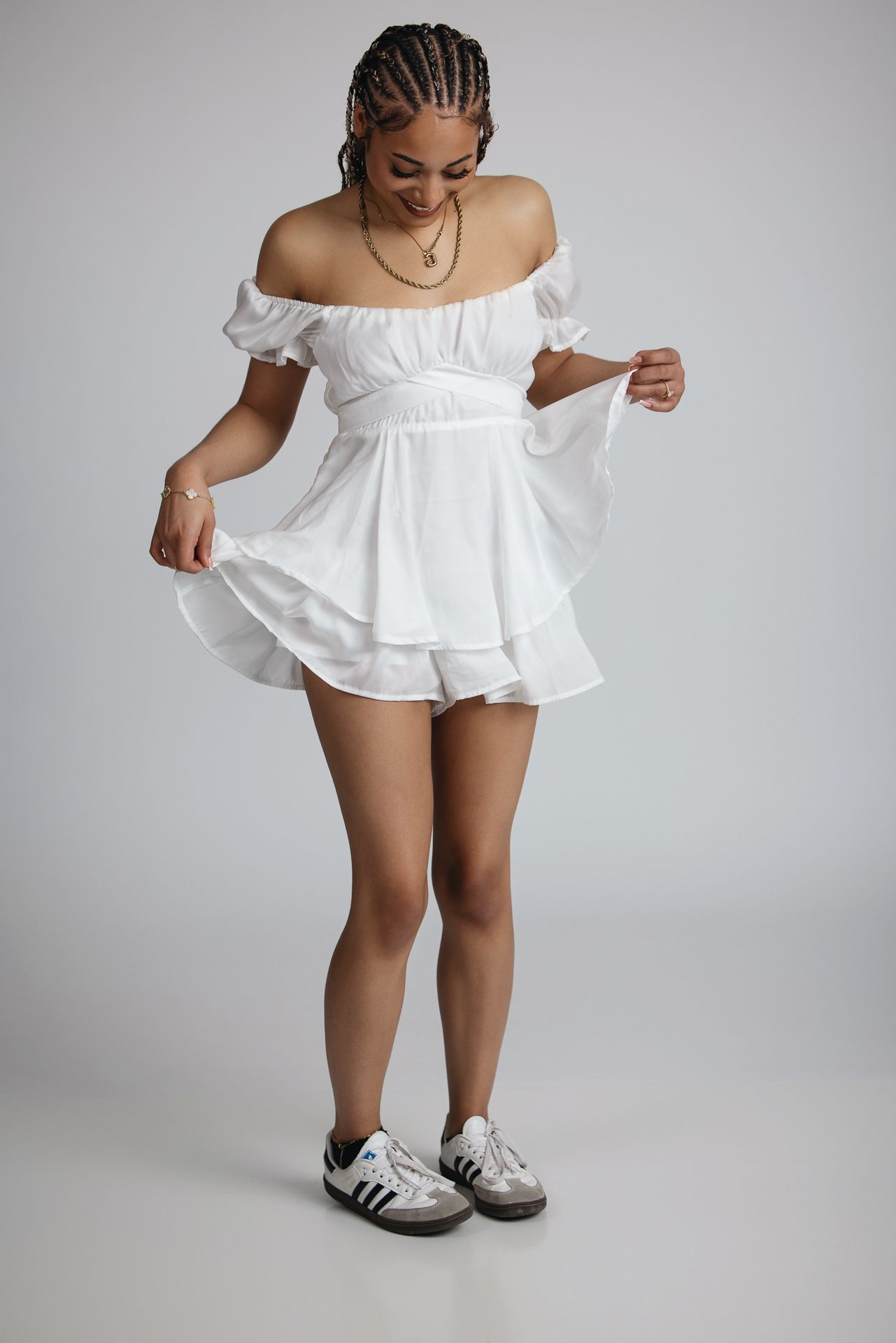 Young woman with long braids in white off-shoulder mini dress and Adidas sneakers in studio portrait.