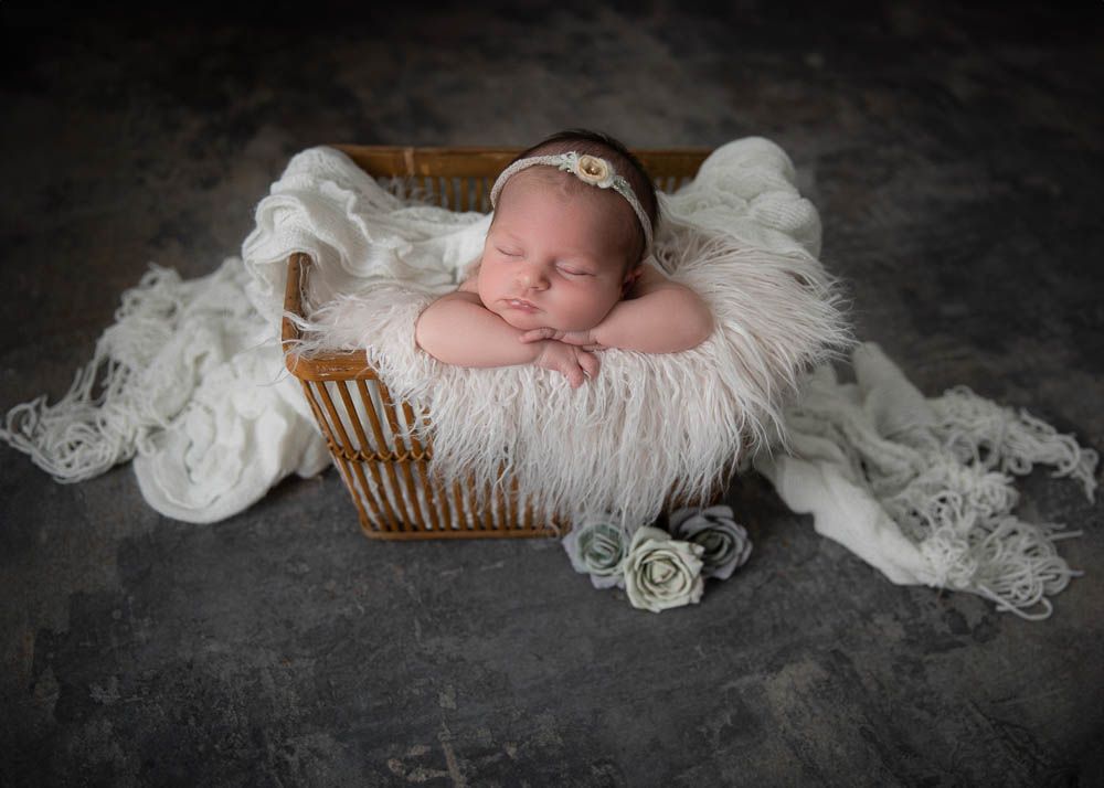 Sleeping newborn baby in white floral headband nestled in a wicker basket with soft white fur.