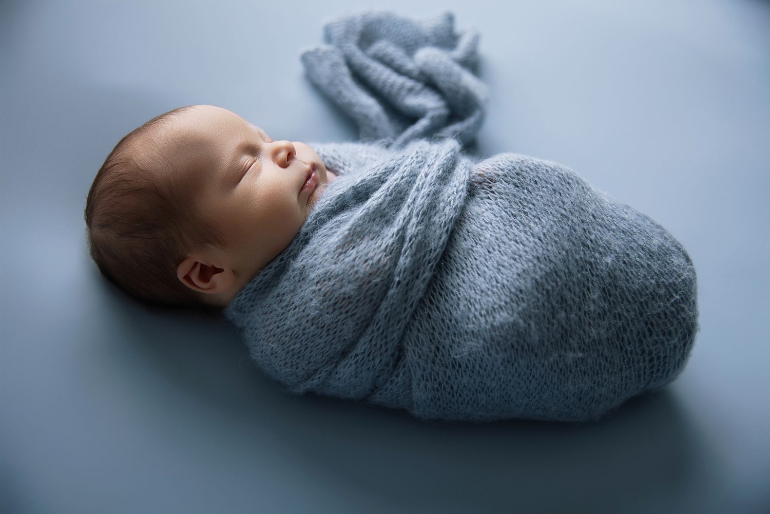 Peaceful newborn baby swaddled in soft blue knit wrap lying on matching blue backdrop.