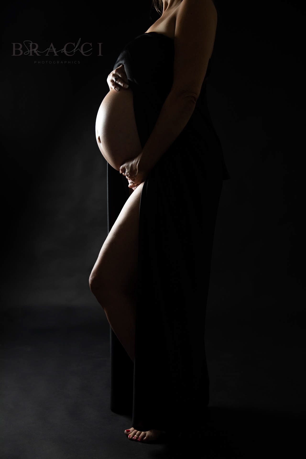 Pregnant woman in black wrap dress holding baby bump in moody dark studio portrait in Bonsall, near Fallbrook, & Temecula