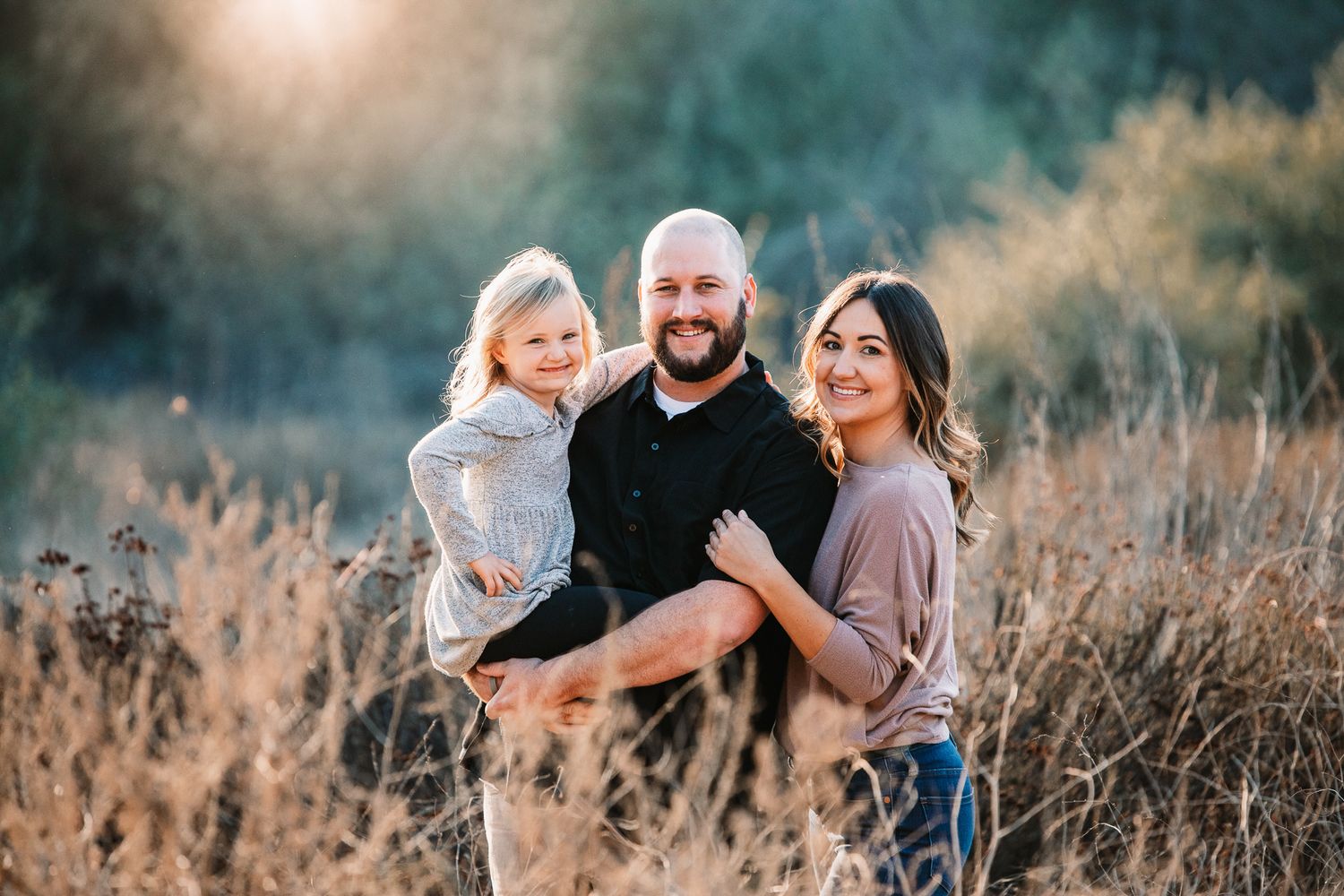 A smiling family of three poses together in a golden sunlit field during a fall photo session.