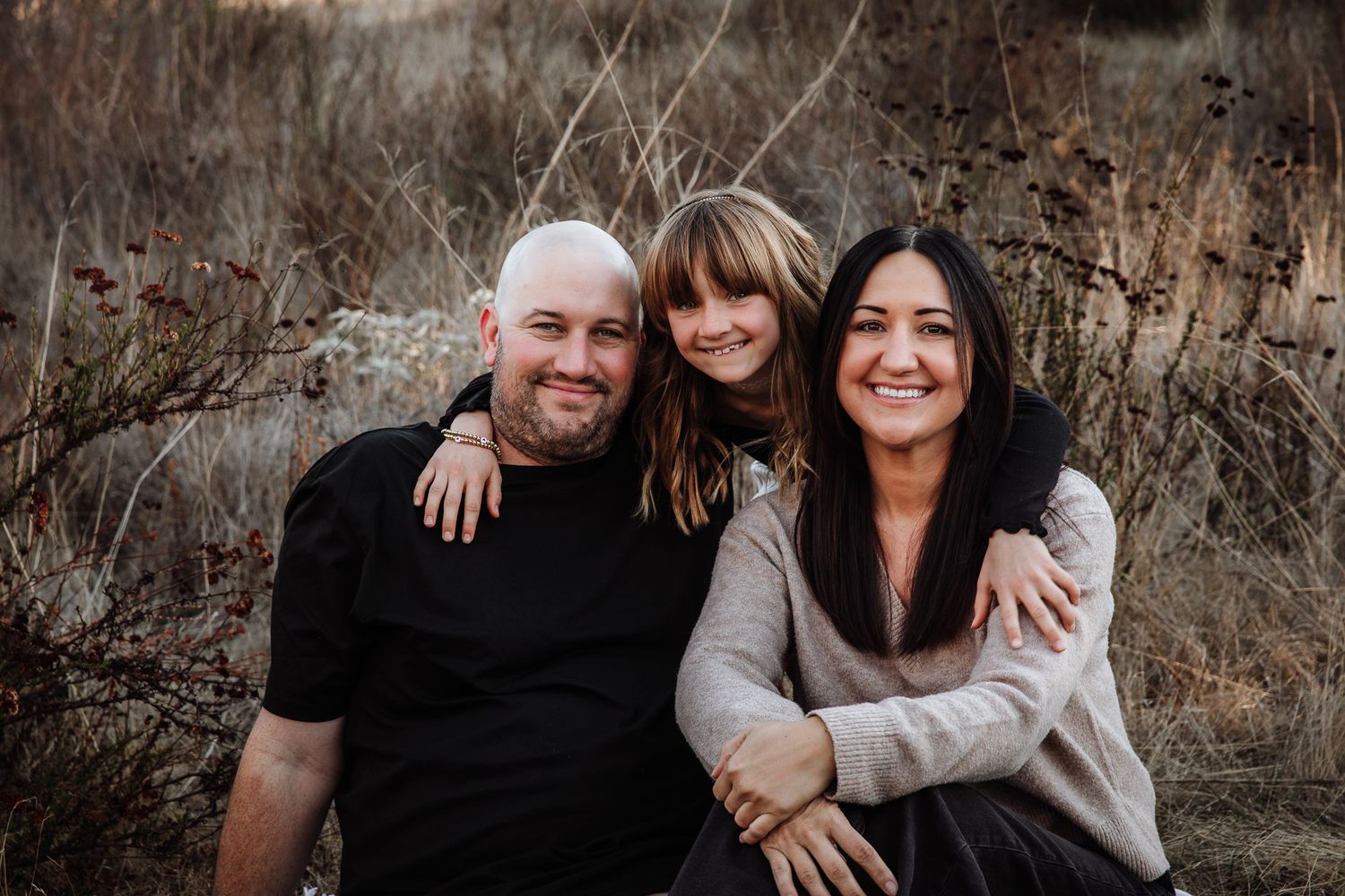 A smiling family of three poses outdoors in a field, with a bald dad, young girl, and brunette mom at a preserve in Bonsall