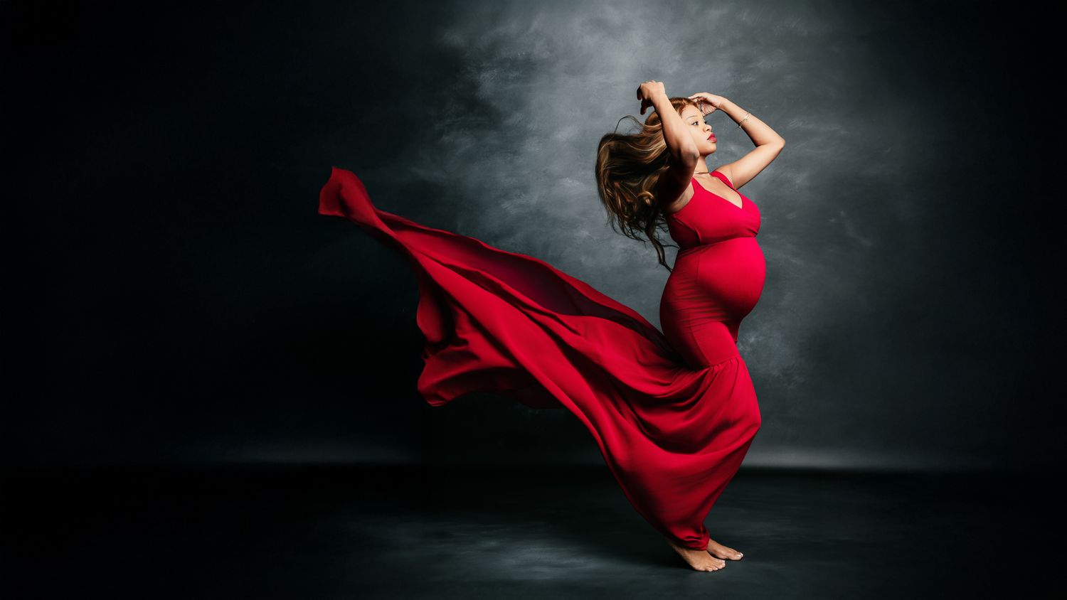 Pregnant woman in flowing red gown dancing dramatically against dark backdrop with fabric billowing around her.