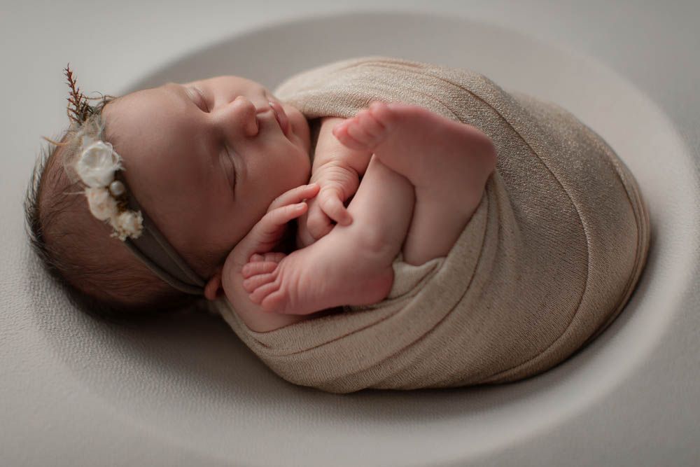 Sleeping newborn wrapped in beige swaddle cloth with delicate white floral headband rests peacefully on neutral backdrop.