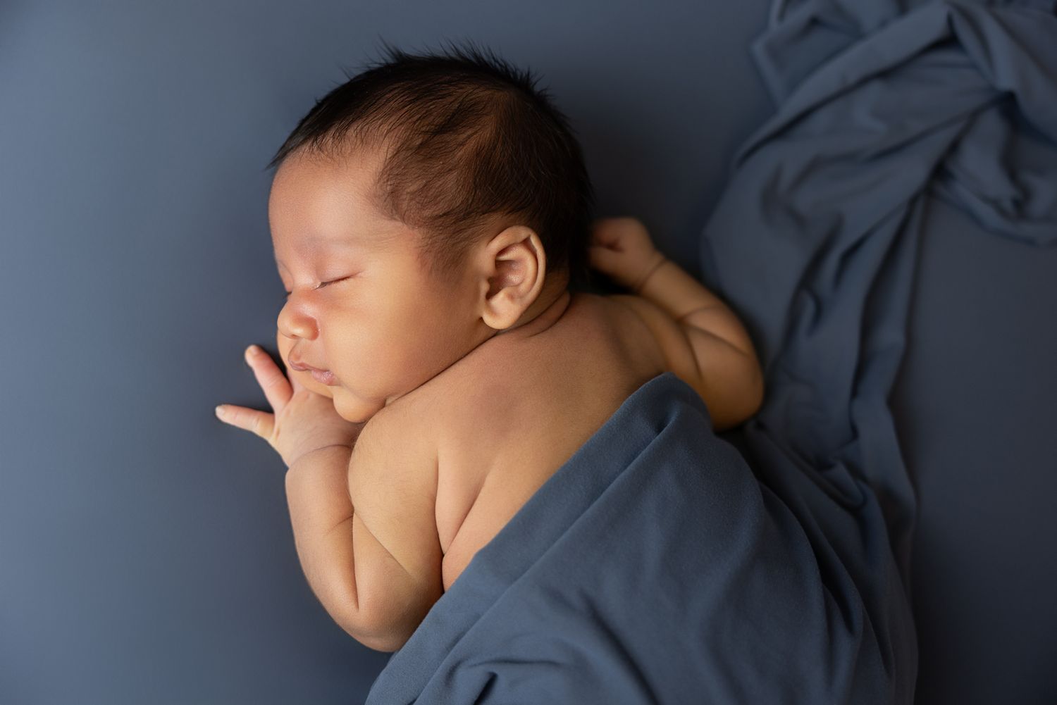 Peaceful sleeping newborn baby lying on a slate blue fabric with hand gently tucked under cheek.