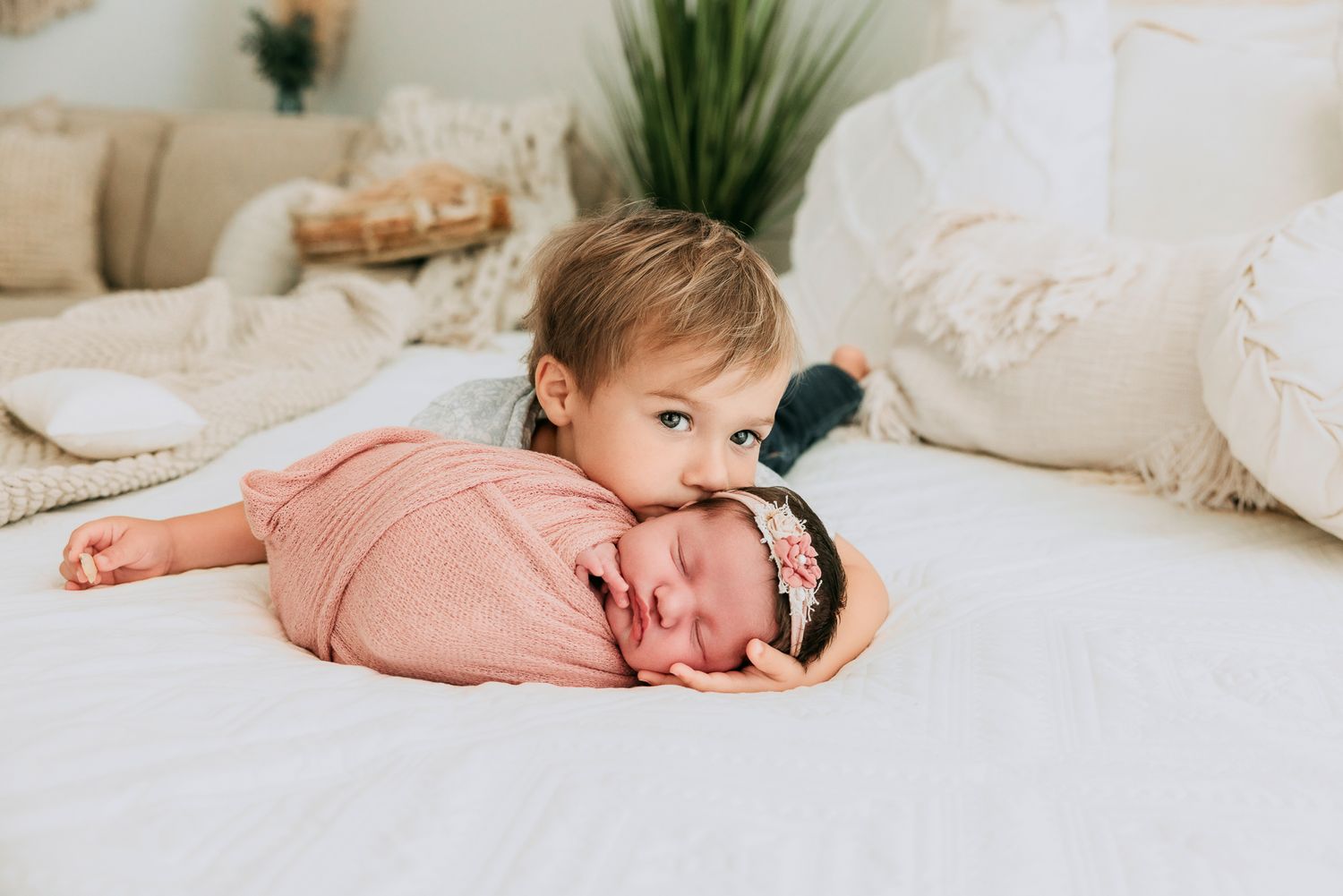 Toddler sibling lovingly poses with swaddled newborn baby in pink on a white bed.