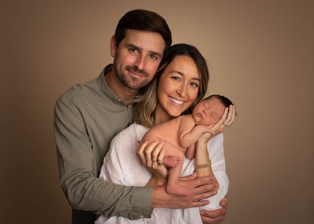 Happy couple smiling and holding their newborn baby wrapped in white cloth against a warm beige background.