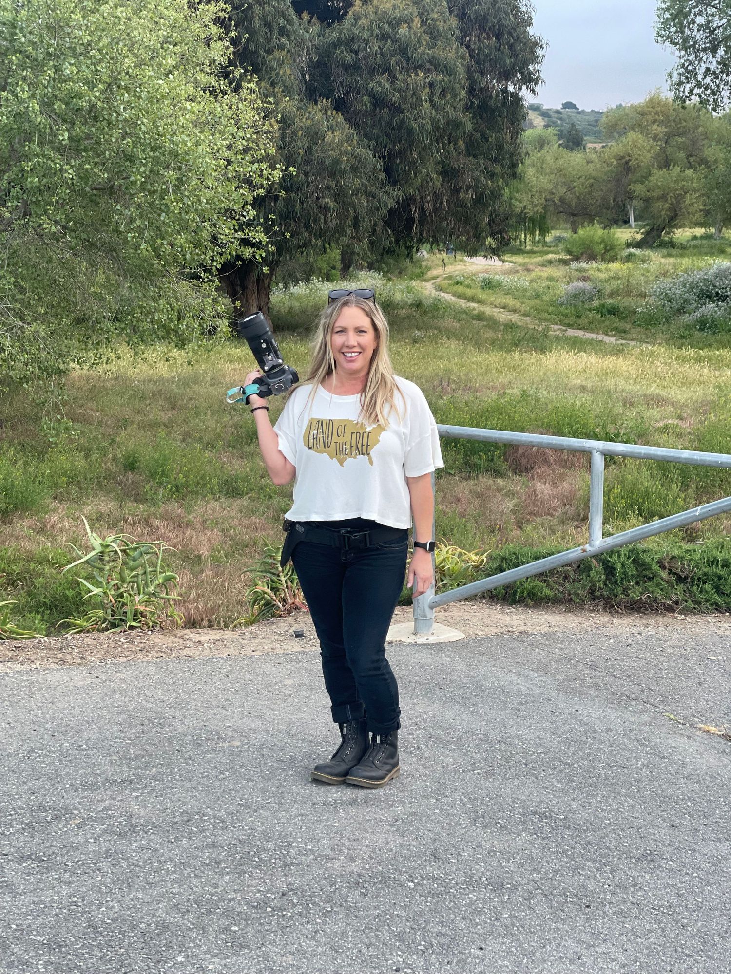 Woman in white graphic tee and black jeans holds camera stabilizer on paved path surrounded by green trees.