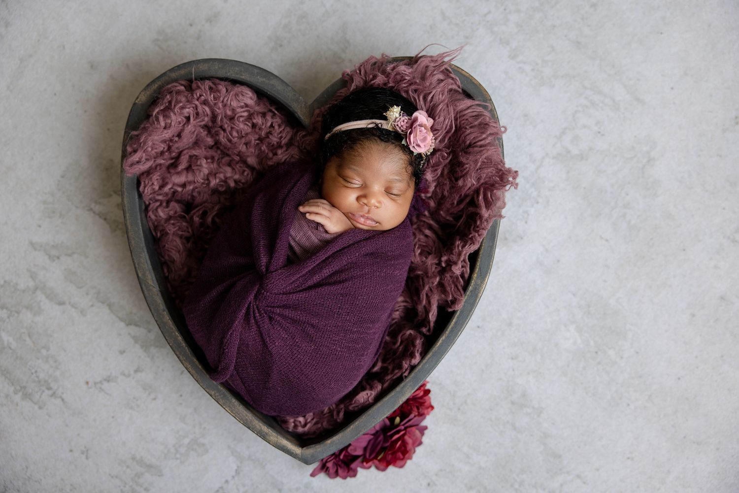 Newborn baby swaddled in purple wrap sleeping in heart-shaped wooden bowl with floral headband.