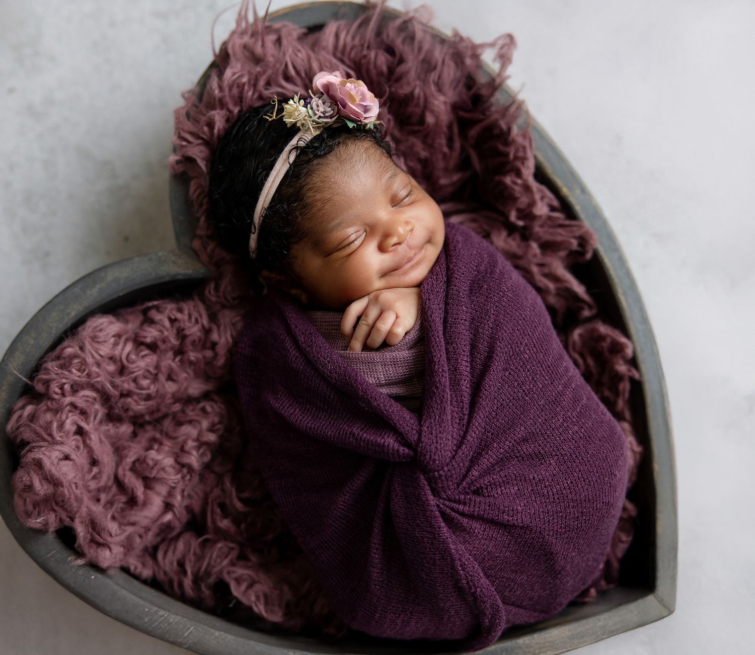 Smiling newborn baby swaddled in purple wrap, wearing floral headband, resting in heart-shaped wooden bowl with mauve fur.