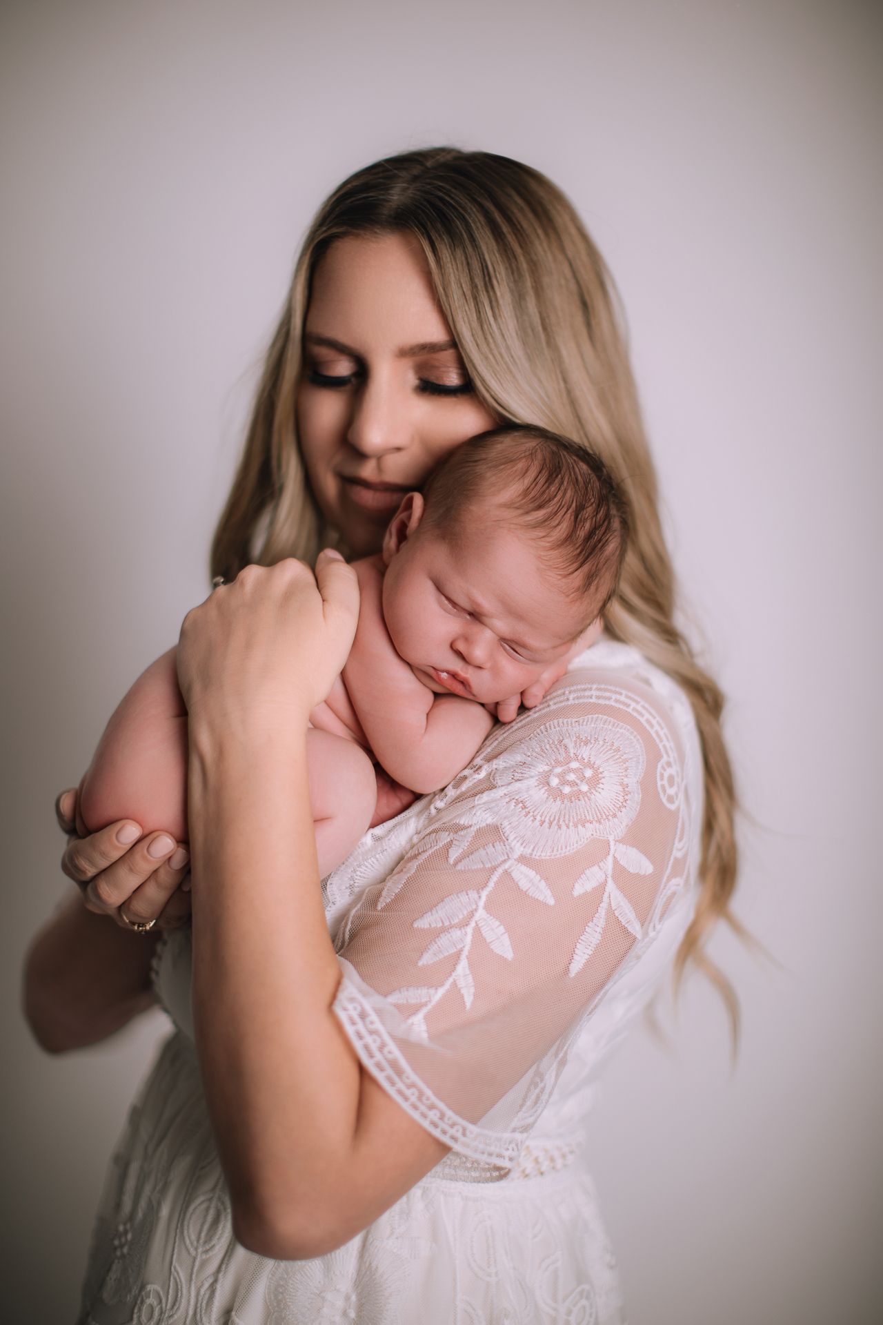 A mother in white lace dress tenderly holds her sleeping newborn baby close to her chest.