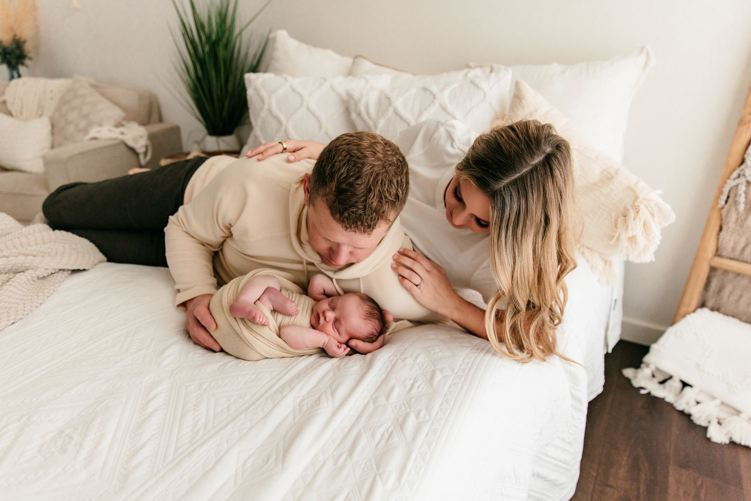 New parents lovingly gaze at their swaddled newborn baby on a white bed during an in-home newborn photo session.