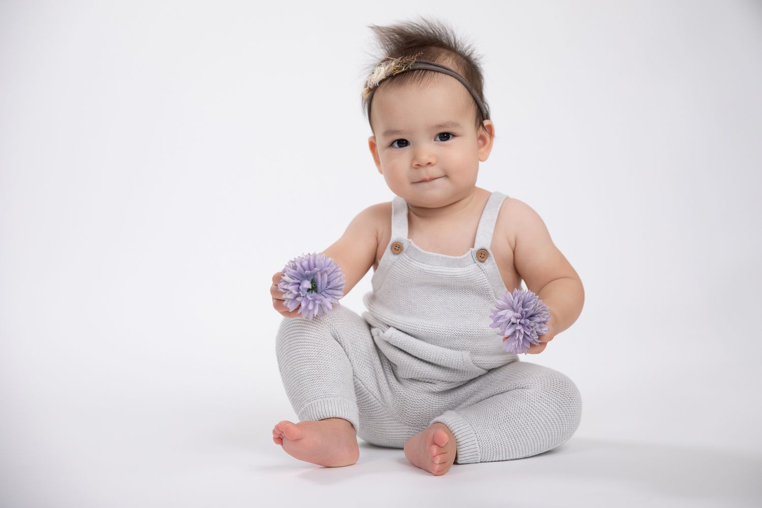 Baby girl in gray overalls holding purple flowers while sitting.
