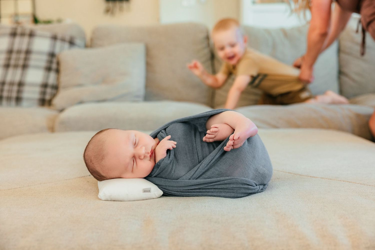 Swaddled newborn sleeping on a white pillow on a couch with an excited toddler in the background.