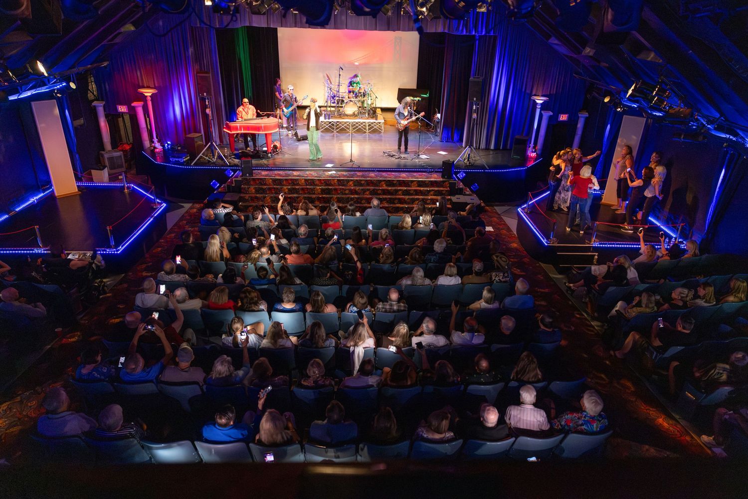 Aerial view of a packed theater audience watching a live band performance on a blue-lit stage.