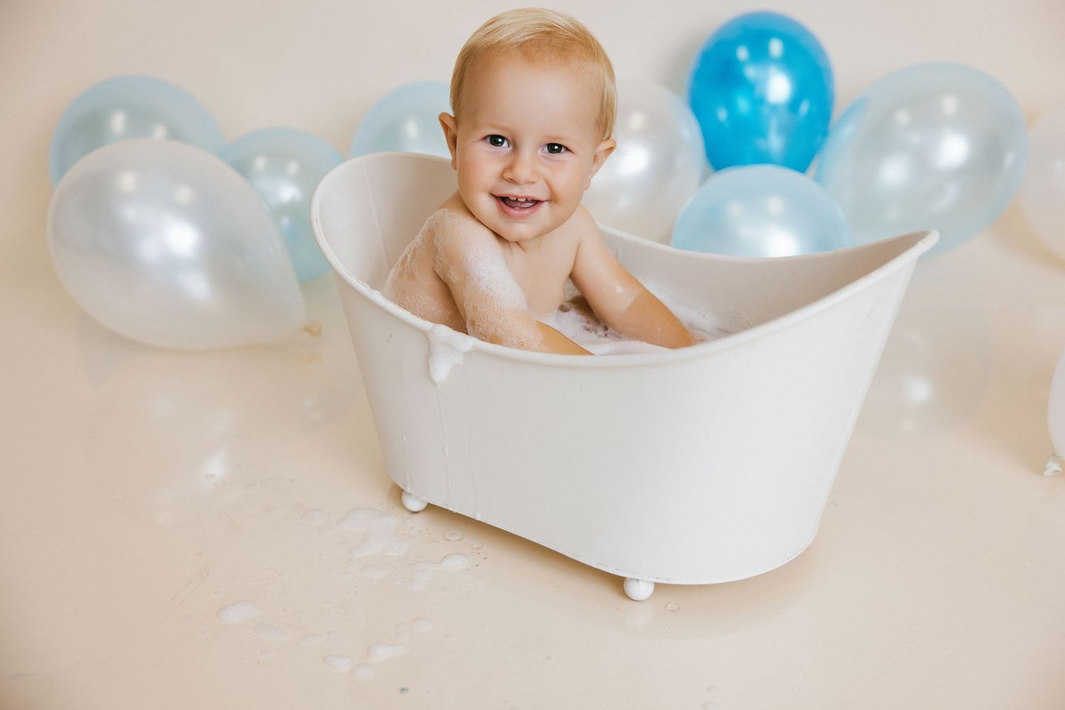 Smiling baby sitting in a small white bathtub surrounded by blue and white balloons.