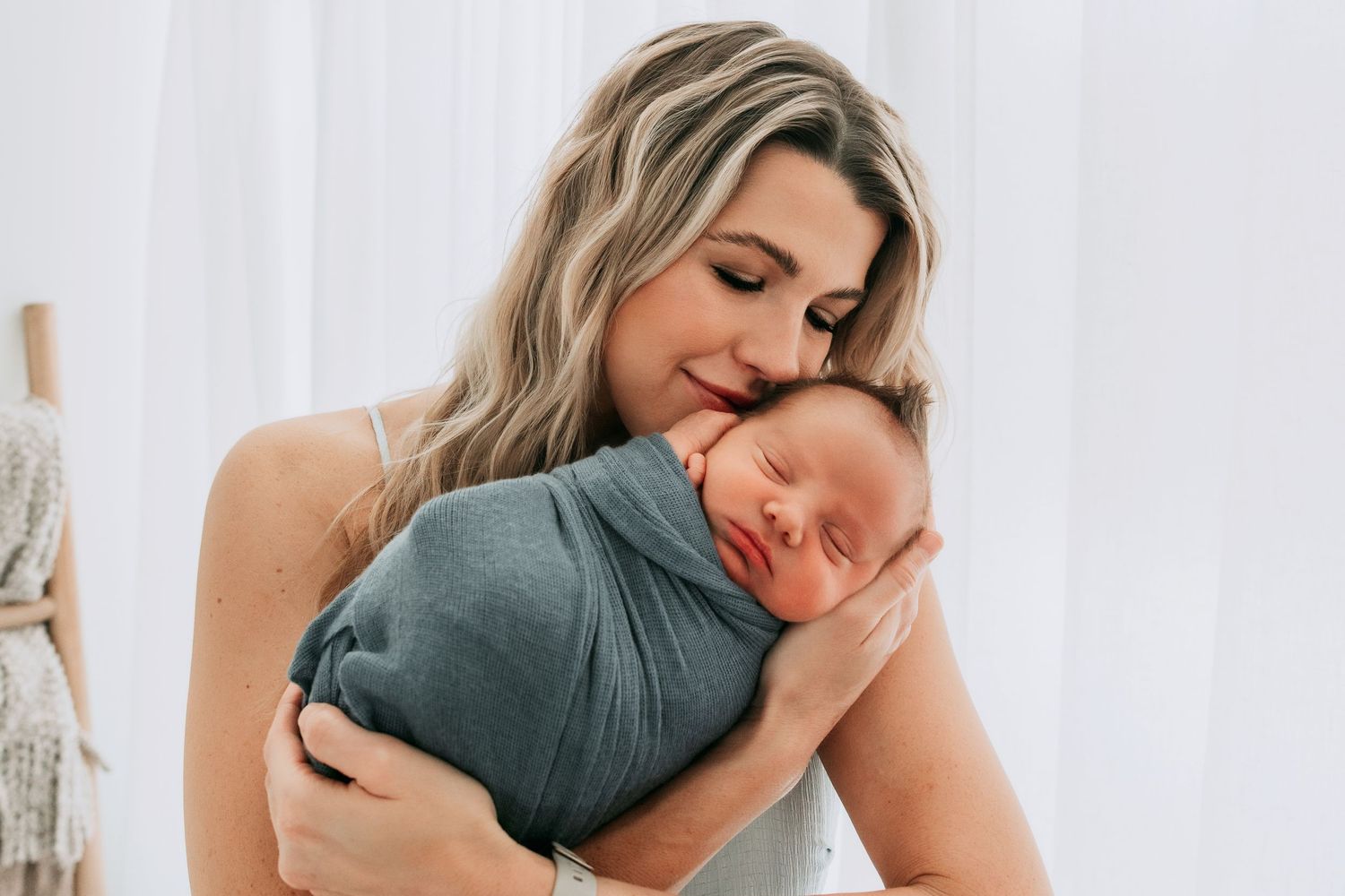 A mother tenderly kisses her swaddled newborn baby wrapped in a teal blanket against a soft white background.