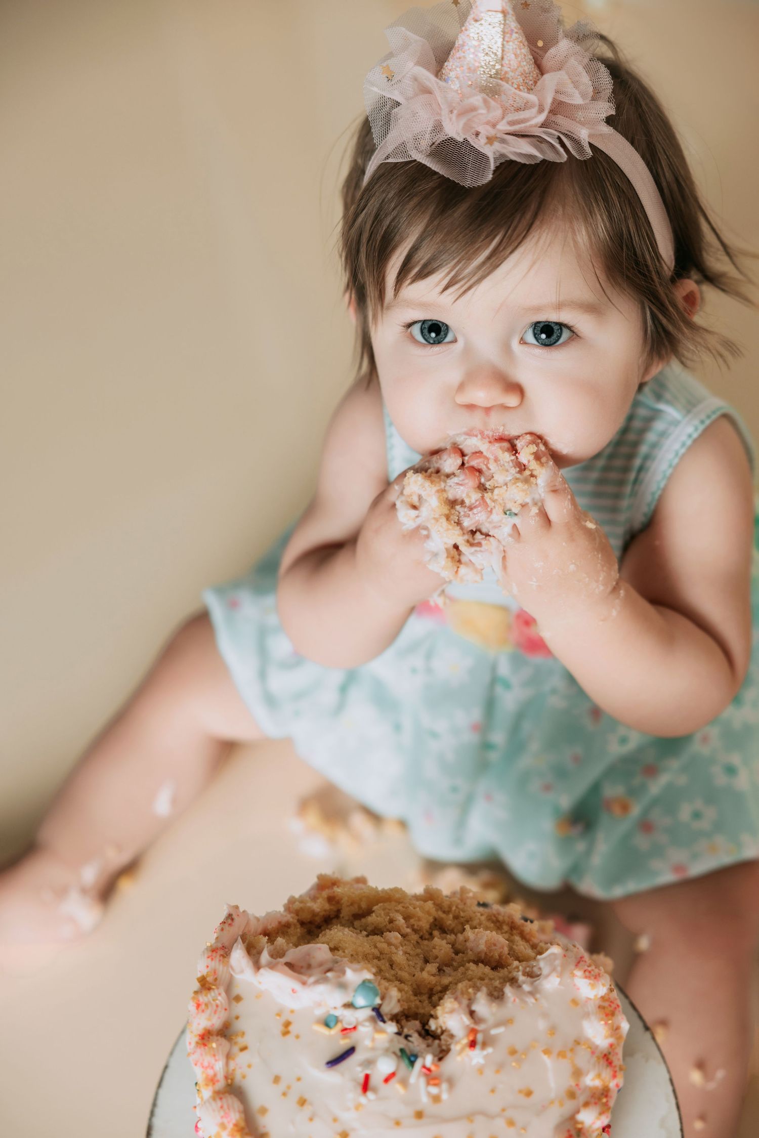 Baby girl in blue dress and party hat smashing her first birthday cake, eating it with her hands.