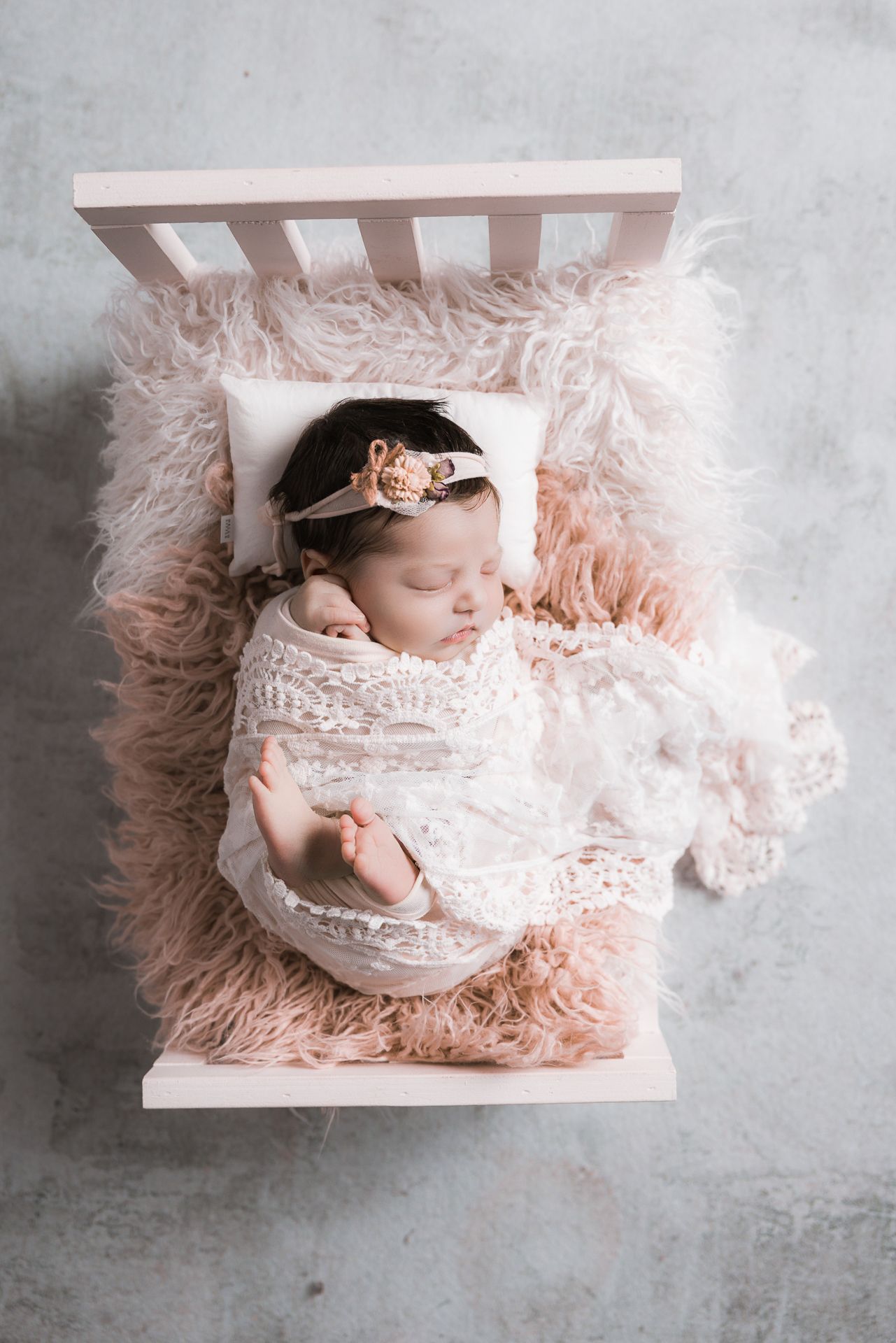 Newborn baby girl swaddled in white lace wrap sleeps in a miniature pink fluffy crib.