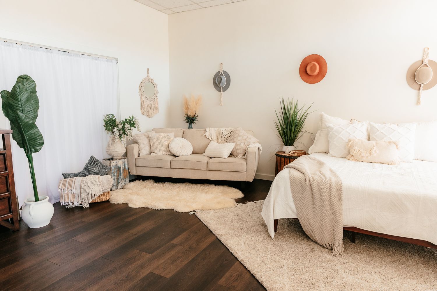 Boho bedroom with beige sofa, white bedding, shag rug, dark hardwood floors, and woven wall decor. in the Bonsall Studio near Temecula