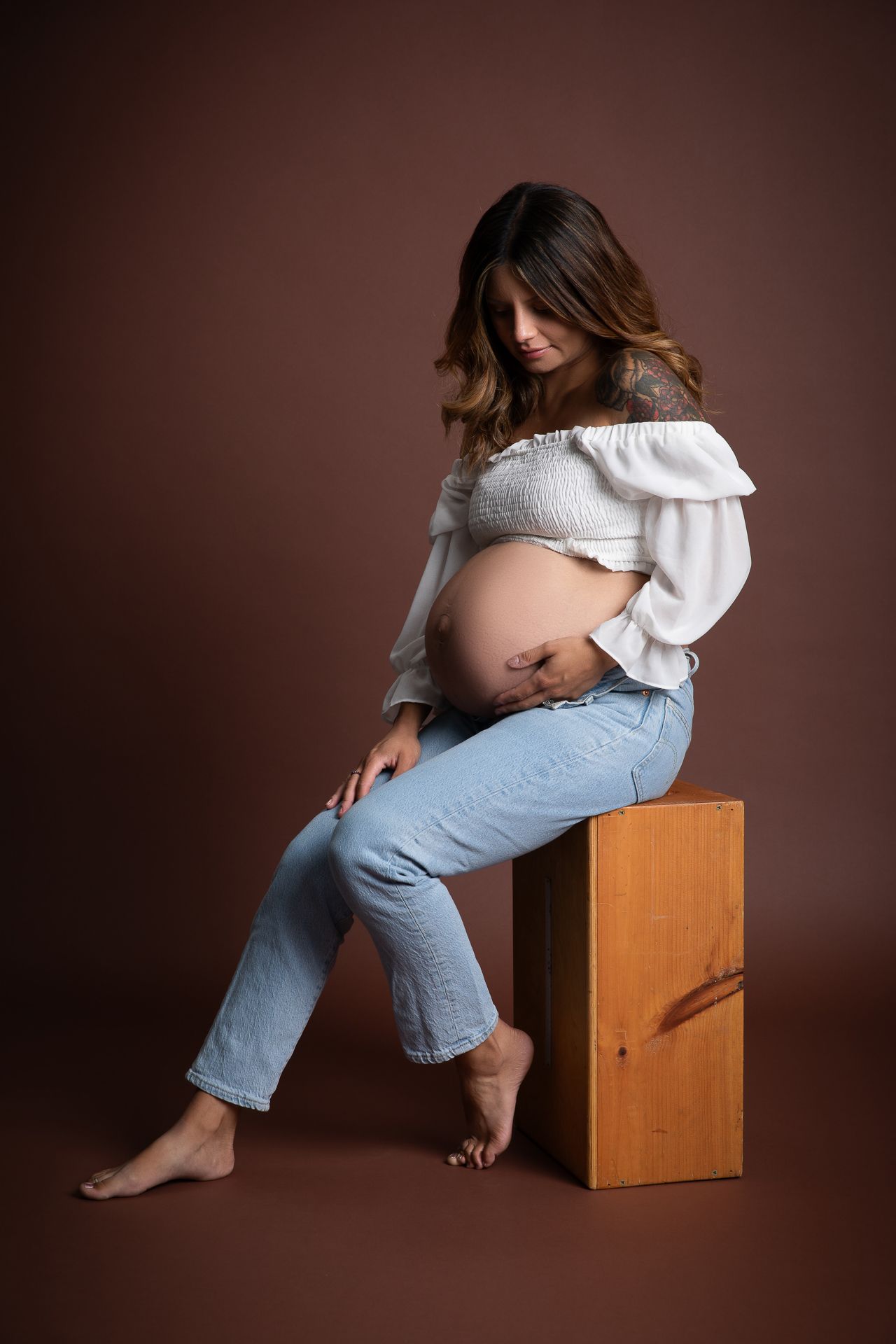 Pregnant woman sitting on wooden box wearing white off-shoulder top and jeans, cradling her baby bump in studio. At the Studio Bonsall, near