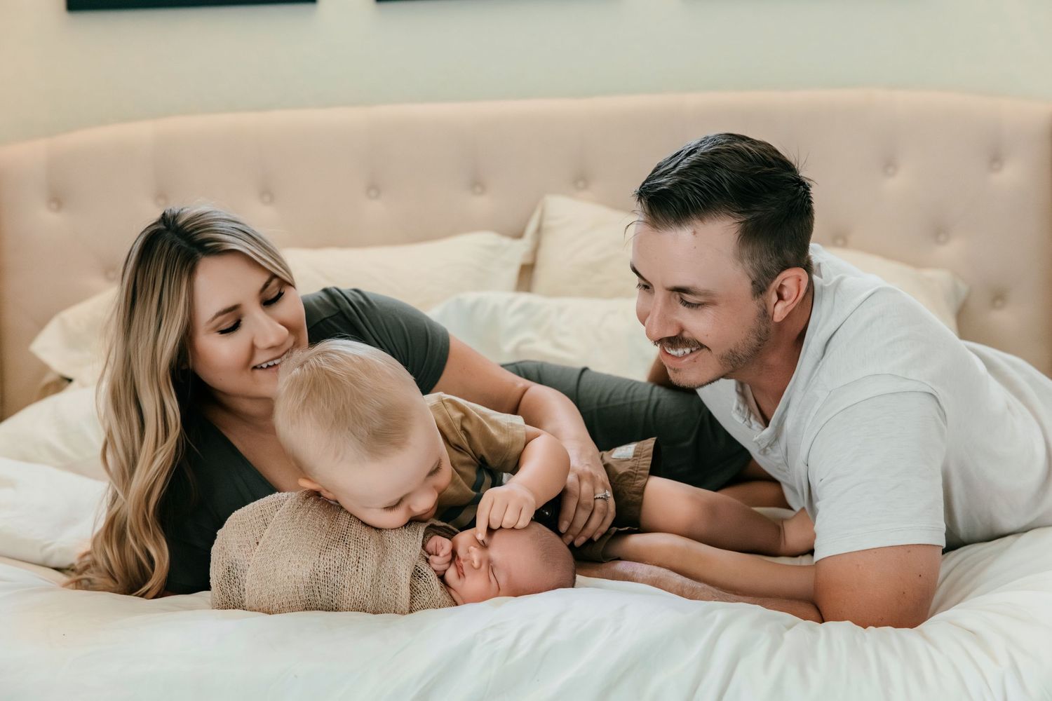 Happy couple smiling at their newborn baby while relaxing together on a white bed in a cozy bedroom.