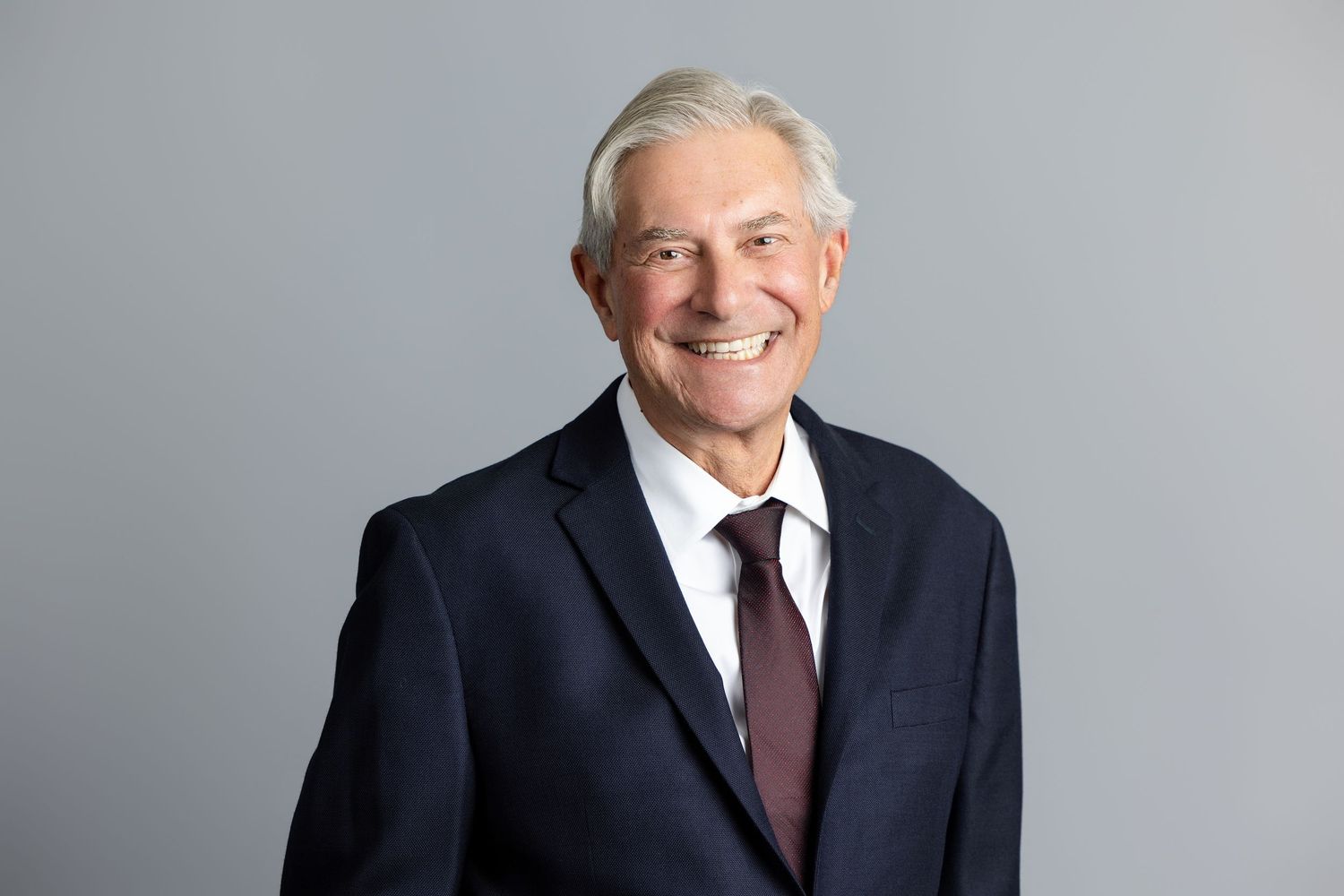 Smiling senior businessman with gray hair wearing a navy suit and burgundy tie against a gray background.