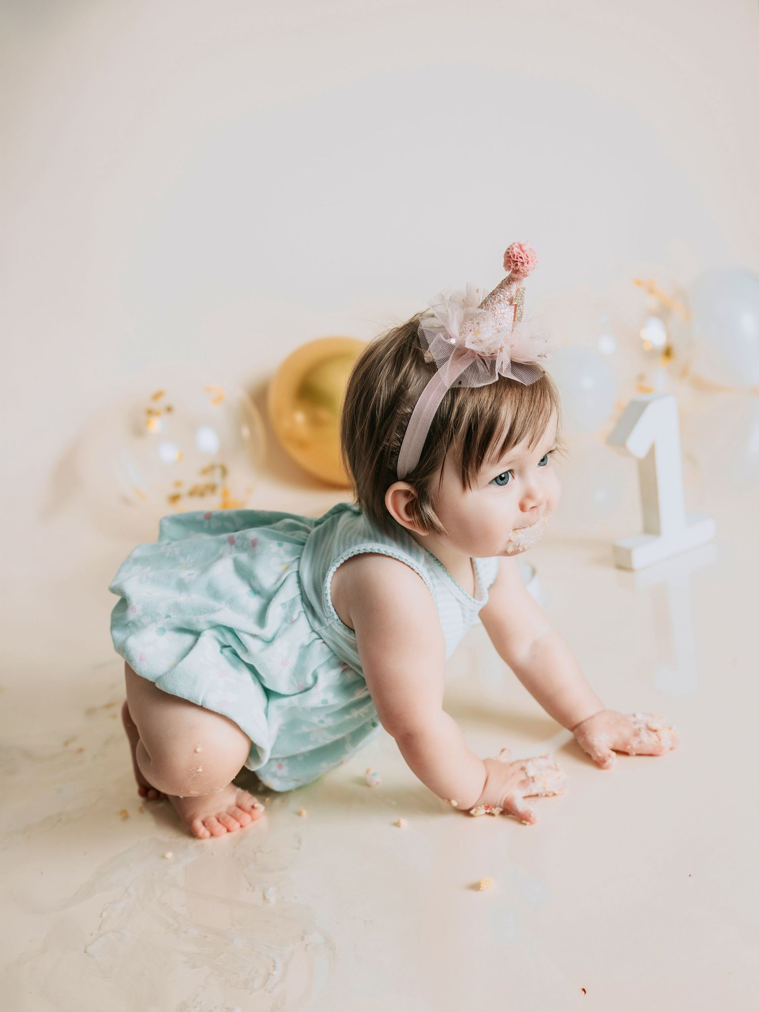 Baby girl in mint romper and party hat crawls during first birthday cake smash photo session.