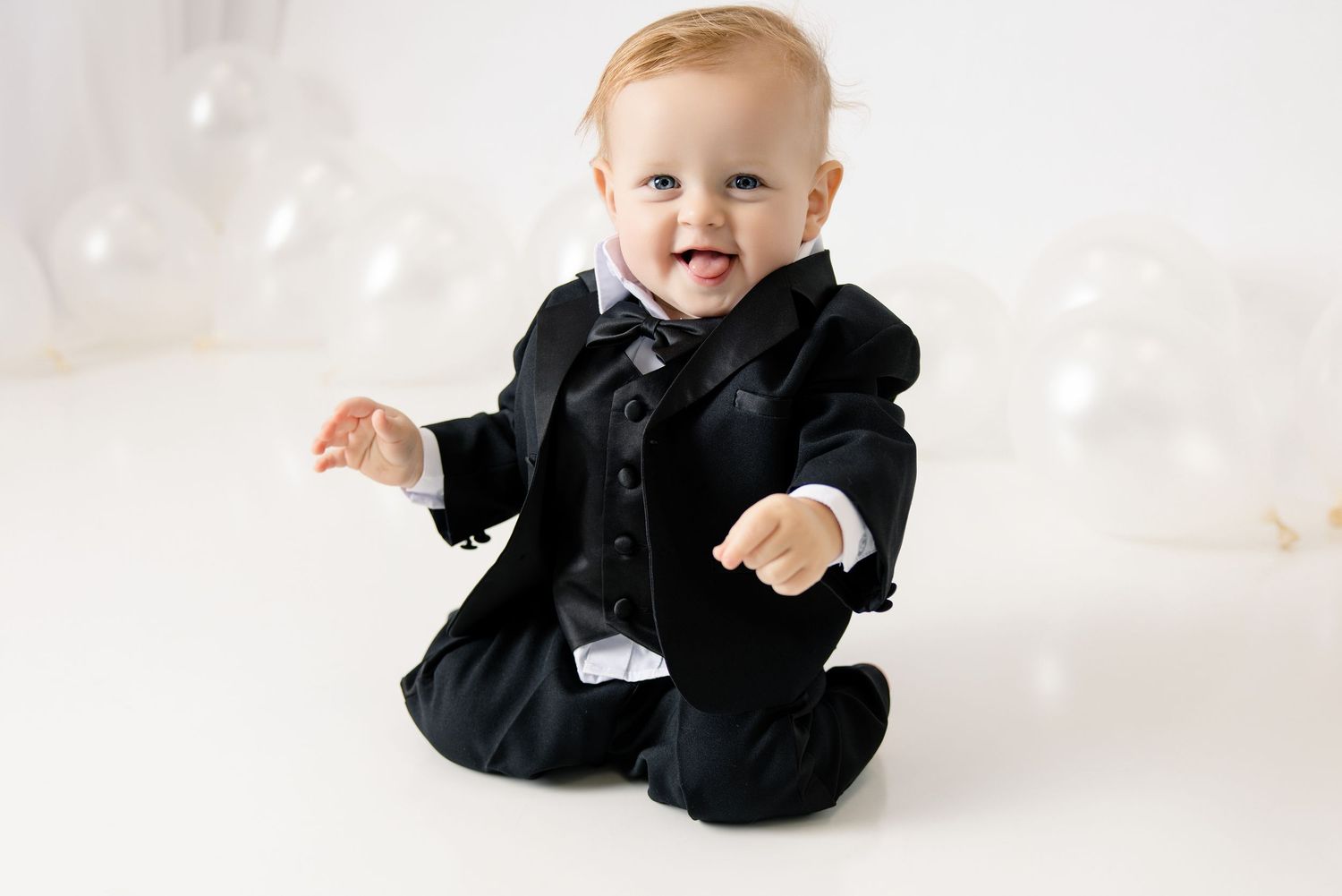 Adorable baby boy in a black tuxedo outfit sitting and smiling against a white background.