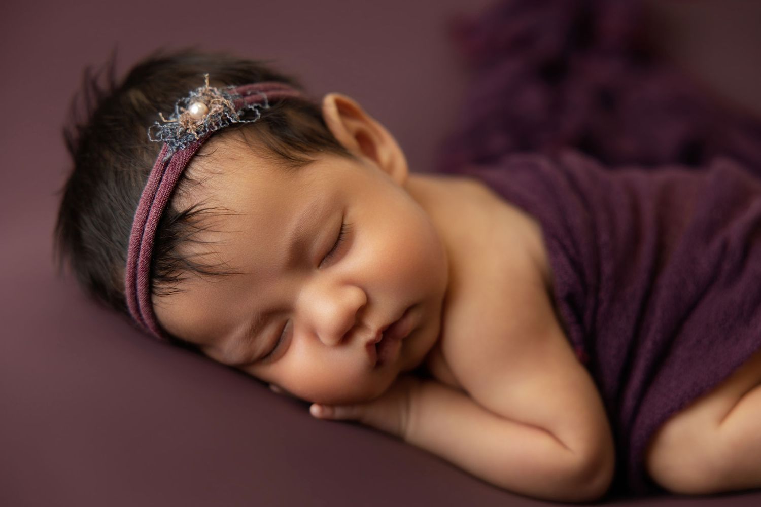 Sleeping newborn baby girl wrapped in purple with a sparkly headband on a mauve background.
