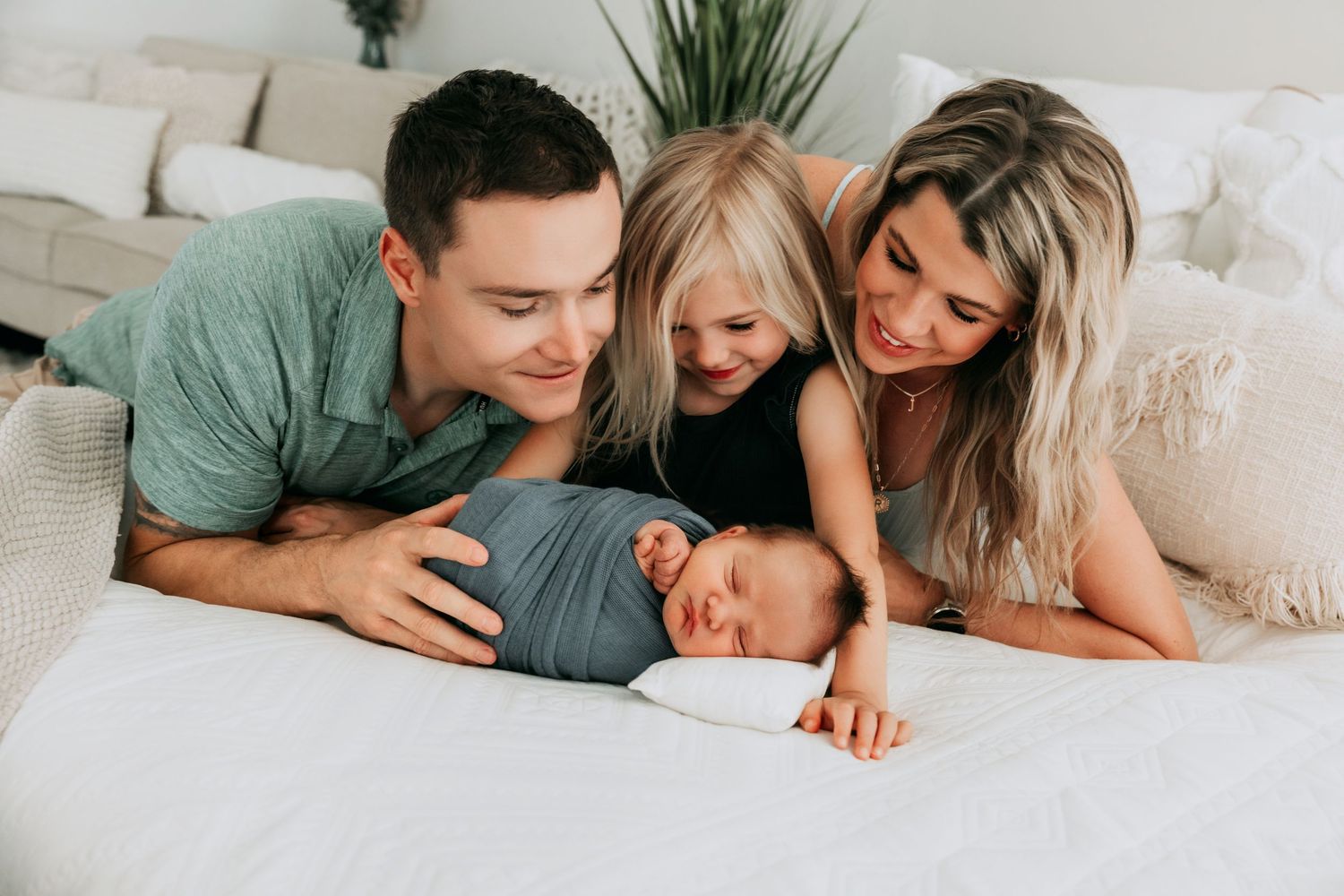 Family of four with parents and toddler lovingly gazing at swaddled newborn baby lying on white bed.