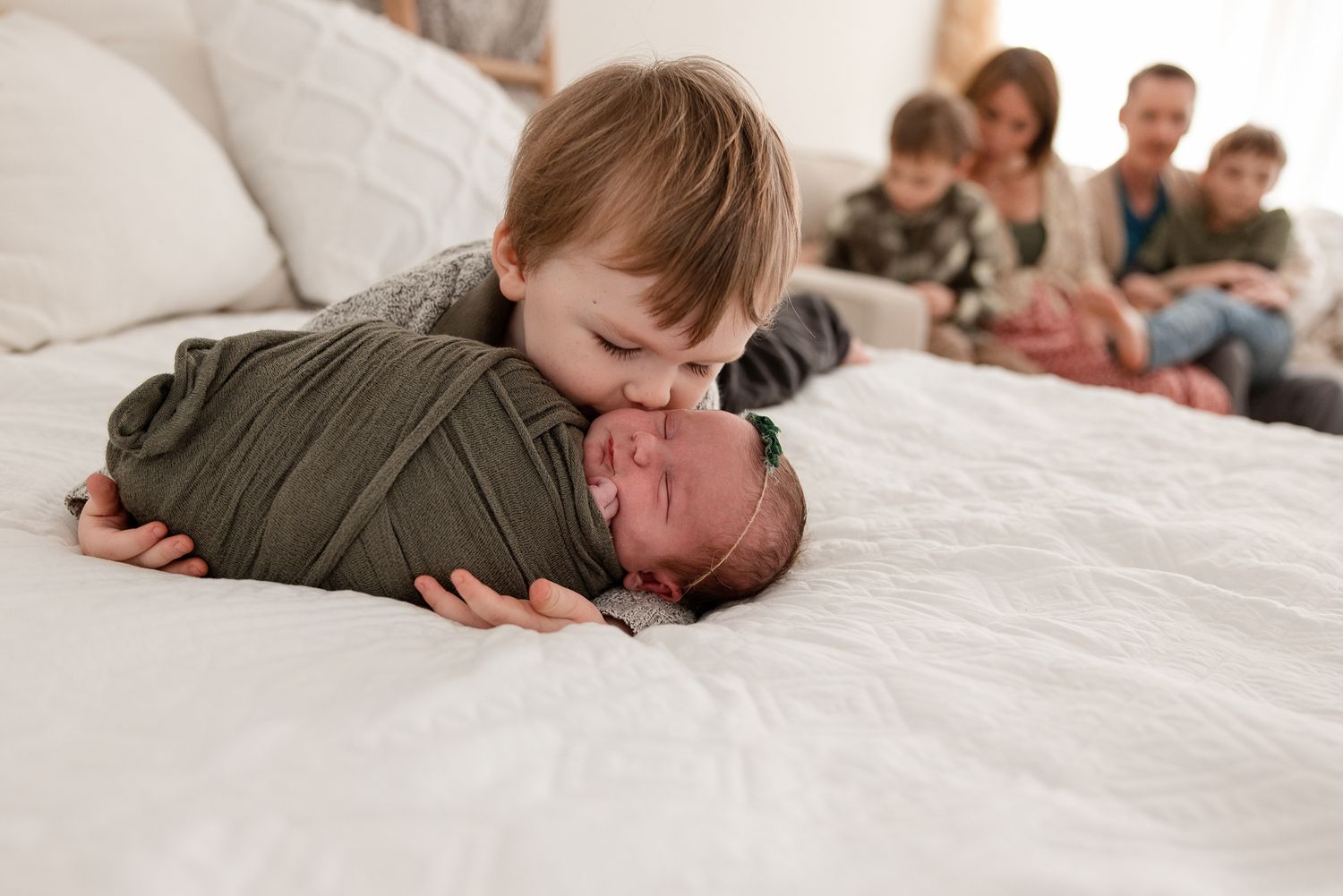 Young boy kissing newborn sibling wrapped in green swaddle on white bed, family watching in background.