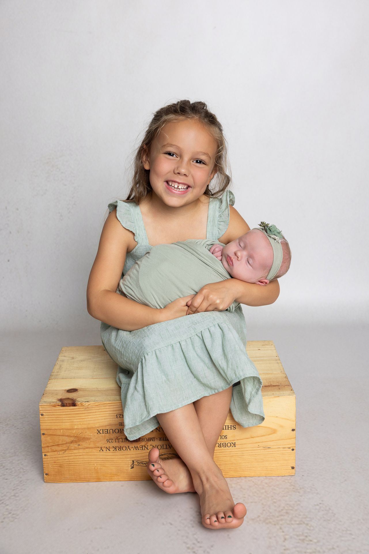 Young girl in mint dress smiles holding sleeping newborn baby sister, both wearing matching outfits, studio portrait.