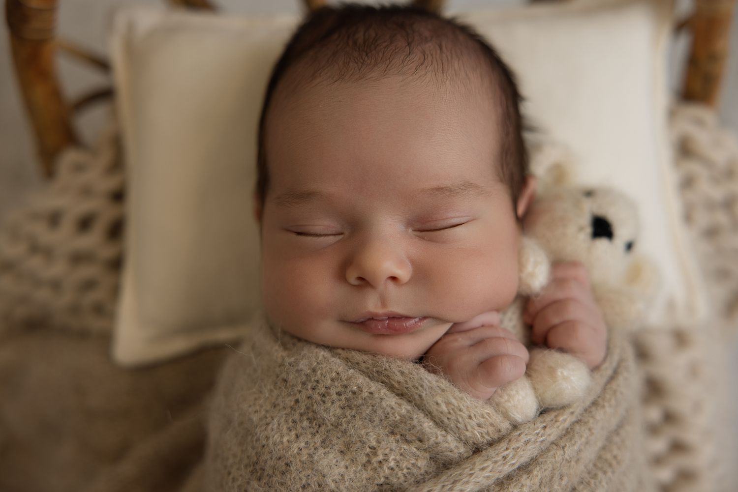 Peaceful newborn baby sleeping swaddled in a soft knit wrap, cuddling a tiny teddy bear on a cozy neutral backdrop.