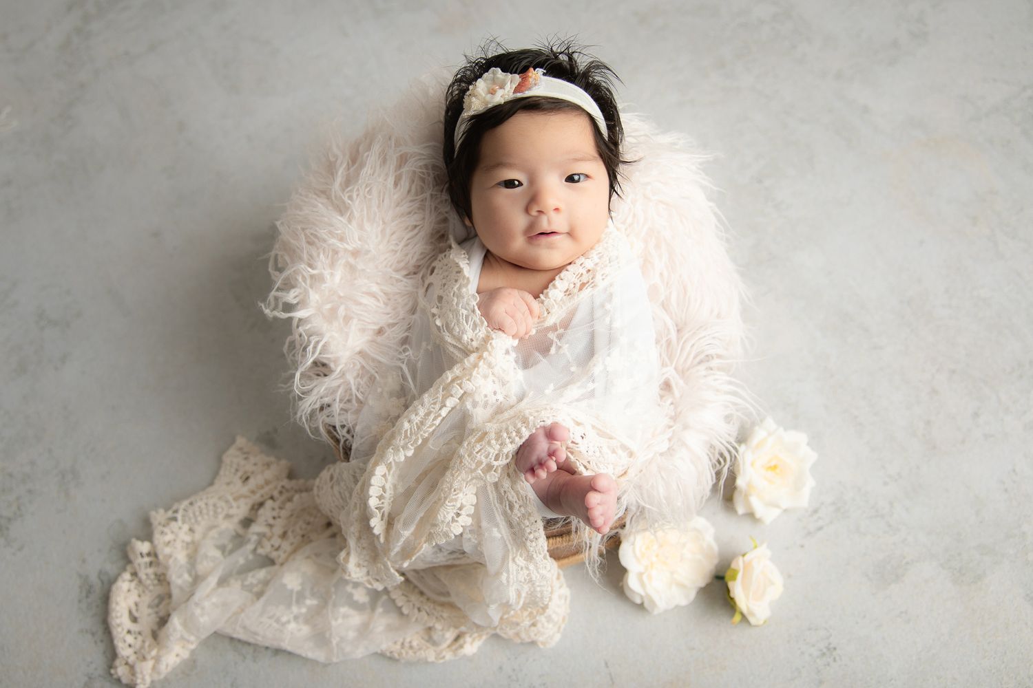 Newborn baby girl in white lace outfit and floral headband lying on fluffy white fur with roses.