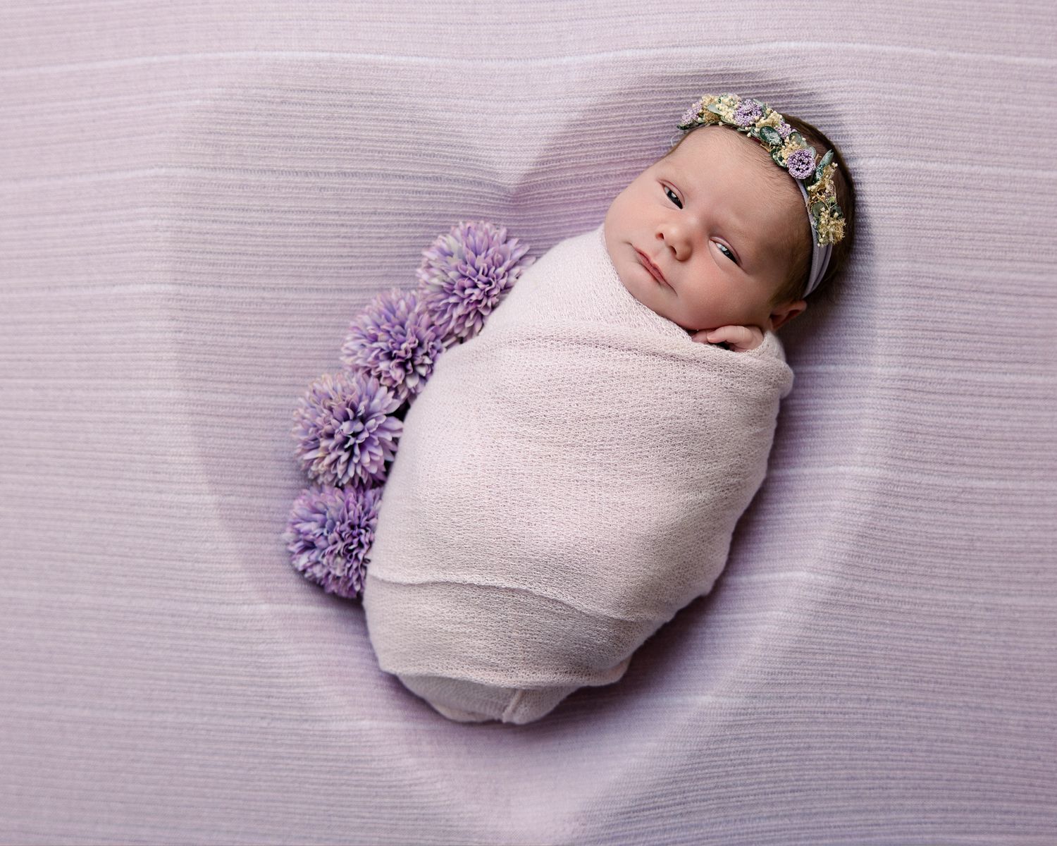 Newborn baby girl swaddled in white wrap, wearing floral headband, sleeping in heart-shaped purple backdrop with lavender pompoms.