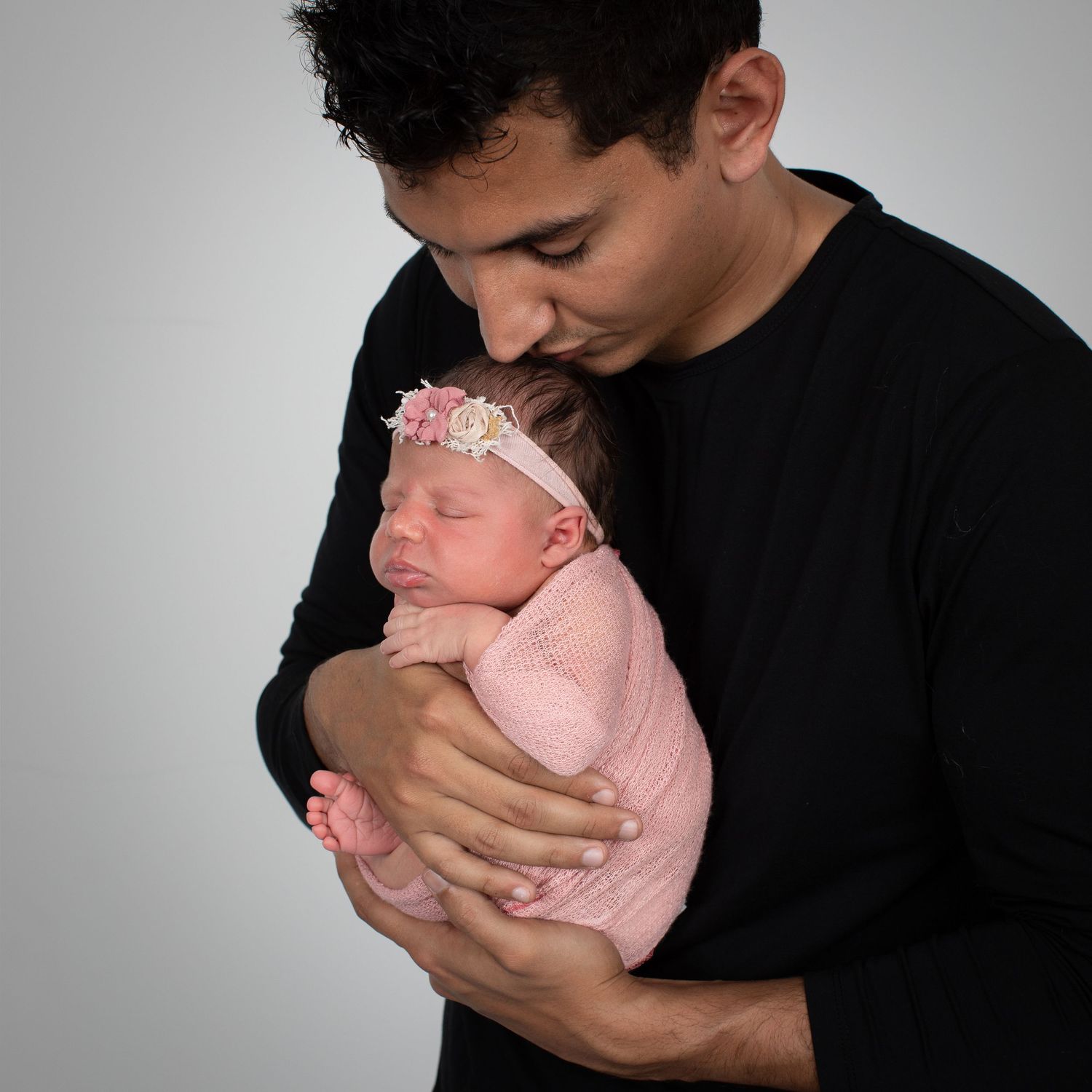 A father in a black shirt tenderly holds his sleeping newborn baby girl wrapped in pink with a floral headband.